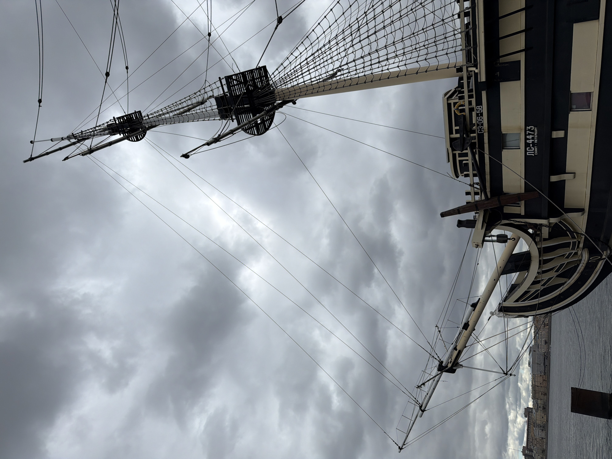 The image shows a tall ship, specifically the Shtandart frigate, in Saint Petersburg, Russia. The ship's black and white hull is visible with the registration number "ЛС-4473 САНКТ-ПЕТЕРБУРГ" and "63-06-58" painted on the side. The ship's masts are adorned with rigging and crow's nests. The sky is overcast with dark clouds. The water of a river and a portion of the city's architecture are seen in the background.