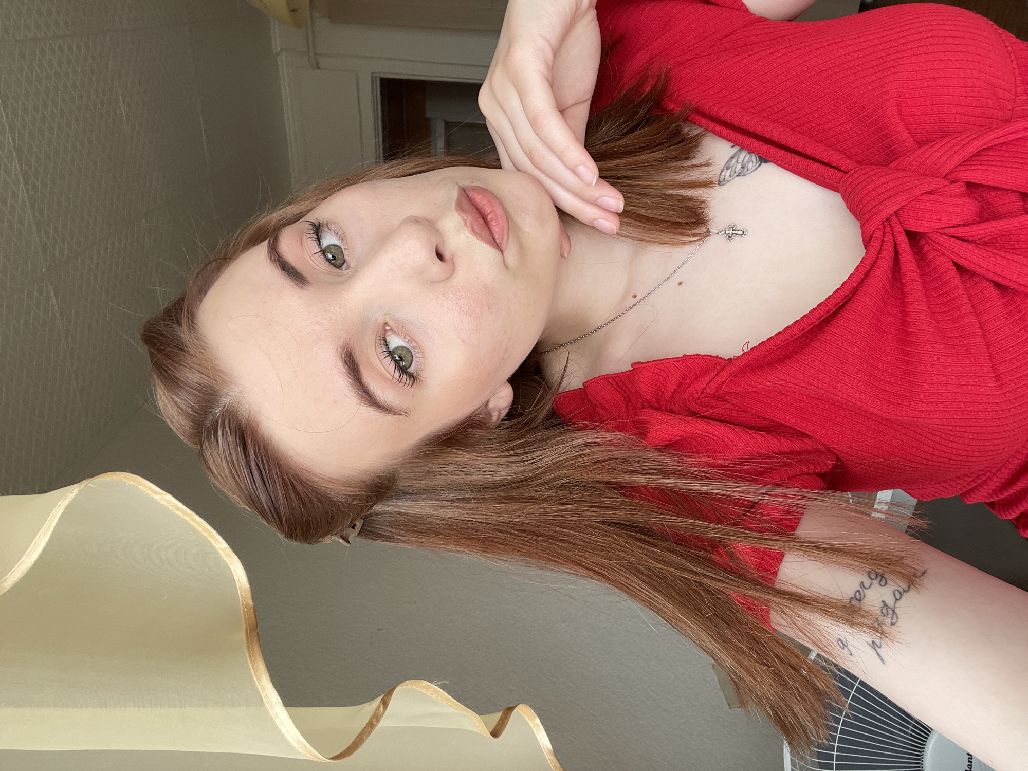 The image features a young woman positioned for a close-up, appearing to recline on her side with her head turned towards the camera. She has medium-length, reddish-brown hair, green eyes with mascara, and defined eyebrows. Her lips are a light pink color, and her fair skin shows several small moles or freckles on her face and chest.
She is dressed in a red, ribbed top featuring a twisted or knotted design across the chest. A slender silver chain with a small cross pendant hangs around her neck. Two tattoos are visible: a faint, possibly winged design on her upper chest/shoulder area, and a script tattoo on her forearm with the partially legible words "sempre" and "paga." Her right hand rests gently near her jawline.
The setting is indoors. A light-colored, possibly textured wall is visible in the background, and a sheer, light-colored fabric, likely a curtain with a golden edge, is present towards the lower right. Part of a white electric fan is also visible in the bottom right corner. The lighting is soft and even, suggesting indoor or diffused natural light. The woman maintains a neutral expression, looking directly at the viewer. The original image is oriented sideways, appearing rotated counter-clockwise.