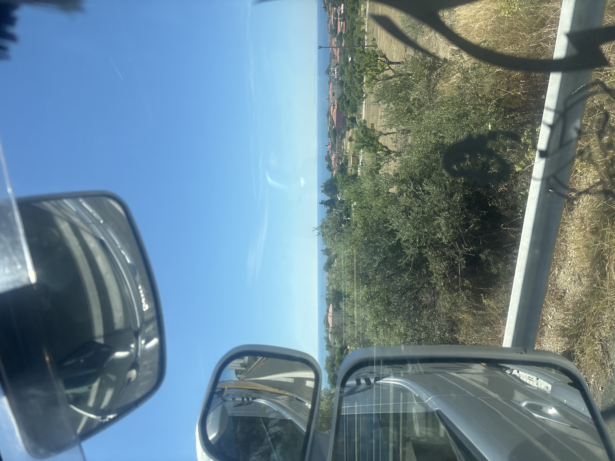 The image, taken from inside a vehicle, shows a bright, sunny day with clear blue skies. The scene is a view of the coast, likely in Makri, Greece, as suggested by the location context. In the foreground, two side-view mirrors are visible, reflecting parts of the vehicle's interior, including a striped pattern on the roof and some metal components.
Beyond the mirrors, a coastal landscape unfolds. A body of water, likely the sea, stretches across the horizon, meeting the sky. Along the coastline, a small village or town is visible, with buildings featuring terracotta-colored roofs nestled amongst lush green trees and vegetation. The terrain appears hilly, with dry grass and olive trees dotting the landscape. A light-colored metal railing or structure runs along the right side of the image, casting a shadow onto the vegetation. The overall impression is one of a warm, pleasant day in a Mediterranean coastal setting. No people are clearly visible, and the focus is on the expansive view and reflections. There is no visible text in the image.