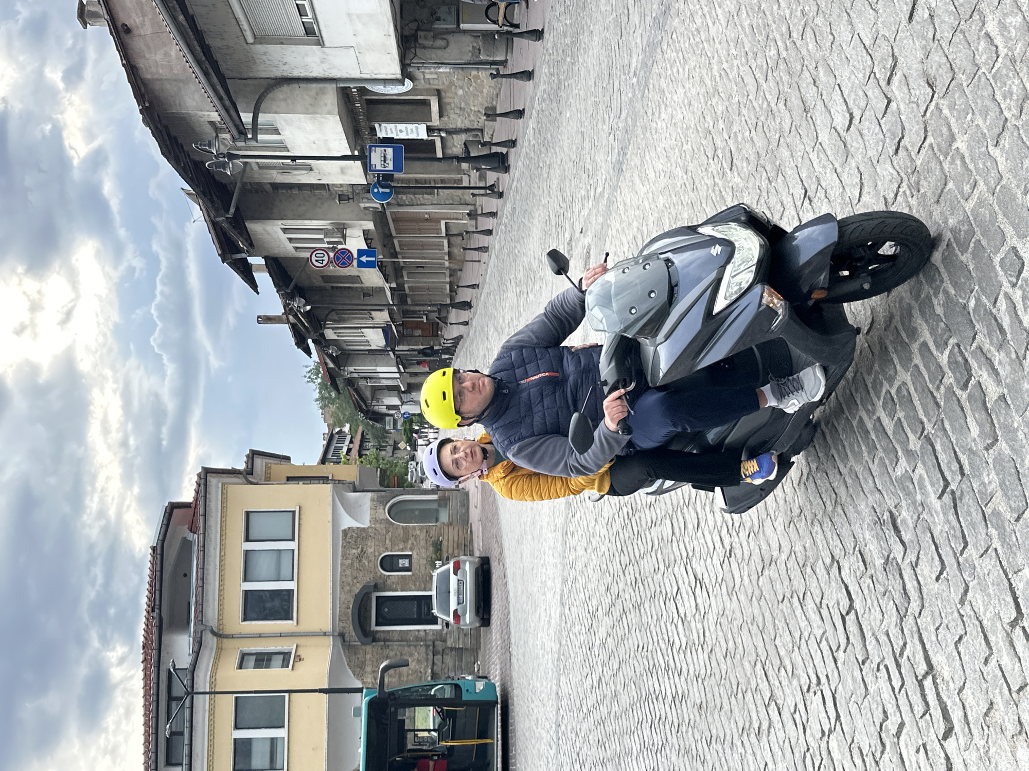 Two individuals, a man and a woman, ride a gray scooter on a cobblestone street in Veliko Tarnovo, Bulgaria. The man wears a yellow helmet and a navy blue jacket, while the woman wears a lilac helmet and a mustard-colored jacket. Behind them are buildings with architectural details and street signs. A bus is visible to the left, and a parked car is further back. The sky is cloudy.