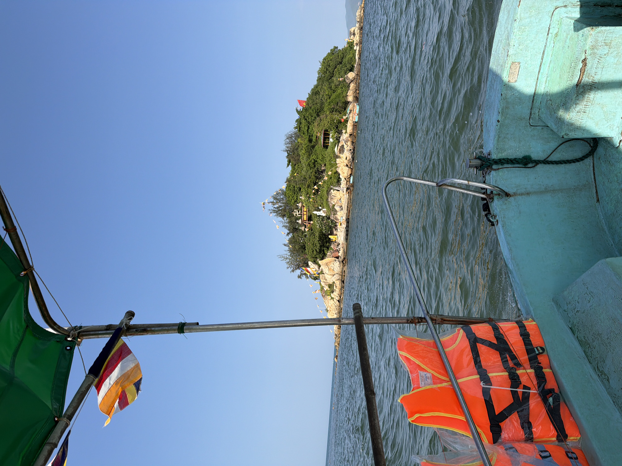 The image captures a view from a boat moving through the ocean towards a small, densely vegetated island. In the foreground, the turquoise-painted edge of the boat is visible, along with two orange life vests sealed in plastic packaging, leaning against a metal railing. A green tarp extends overhead, supported by metal poles, and a colorful striped flag flutters from one of these poles.
The island in the distance is rocky with numerous trees and foliage covering its surface. Several structures, including what appears to be a pagoda or temple with tiered roofs and smaller buildings, are visible on the island. Various flags and banners are also dotted across the island's landscape, adding to its visual complexity. The water is a deep blue-green, with small waves indicating movement. The sky is clear and bright blue.
This scene is consistent with the appearance of Hon Chong (Chong Promontory) in Nha Trang, Vietnam. Hon Chong is a cluster of large rocks on the coast of Nha Trang, often with small pagodas or Buddhist shrines situated among them. The presence of the boat and life vests suggests a tourist excursion or transport to the island.