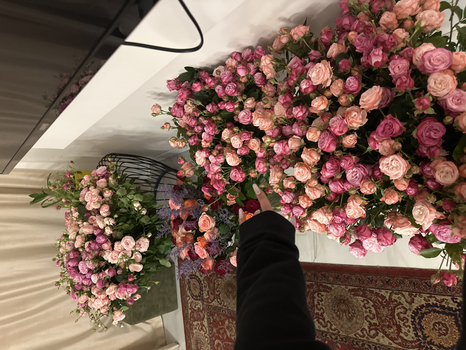An indoor scene in Yerevan, Armenia, displays numerous cut flowers arranged in significant quantities. The lower right and central foreground are dominated by large volumes of cut spray roses in various shades of pink, salmon, and fuchsia. On the left side of the frame, another substantial collection of similar spray roses rests atop a dark green upholstered cube, positioned next to a black wire basket. Between these two primary floral masses, a third arrangement features larger cut roses in red, orange, and pink hues, mixed with purple floral accents. A person's left arm, covered by a dark, long-sleeved garment, and a hand with light-colored manicured fingernails, extends into the frame from the left, positioned in proximity to the central flower cluster. The flooring consists of a traditional red and cream patterned rug on the left and white marble or light-colored tile in the center. A light-colored curtain is visible in the upper left background. A black, flat-screen television is partially visible, mounted on a white wall in the upper right quadrant of the image, with a black cable hanging from it.