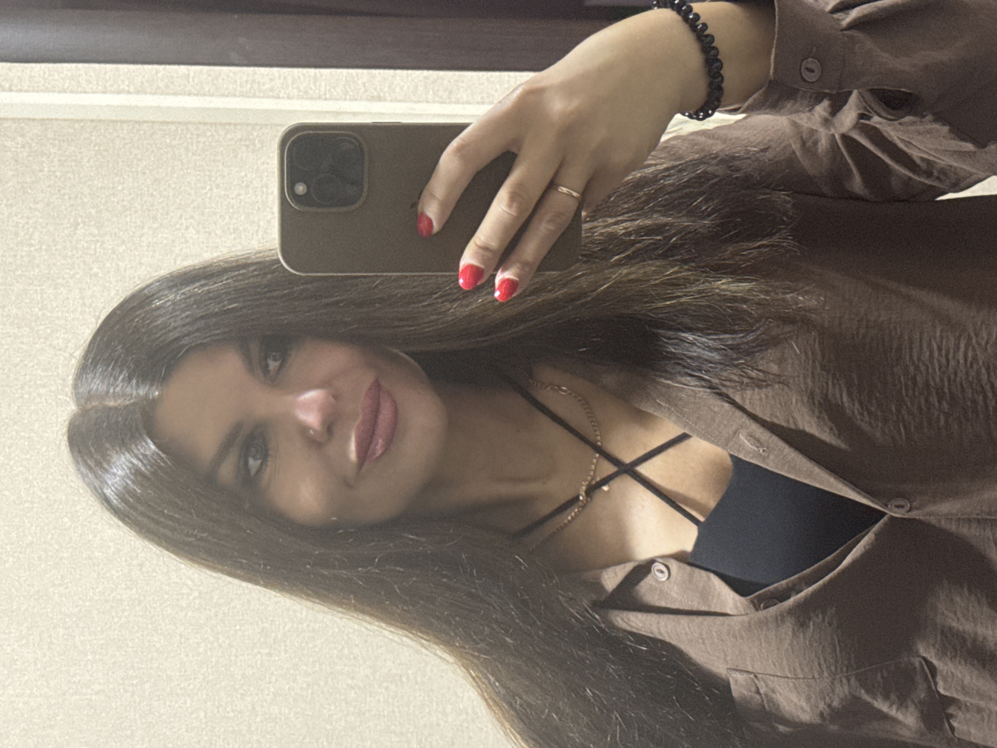 The image shows a young woman taking a mirror selfie. She has long, dark brown hair and is wearing a brown, long-sleeved button-down shirt over a black camisole with crisscross straps. Her fingernails are painted bright red, and she is wearing a thin gold ring on her left hand and a beaded bracelet on her right wrist. The background appears to be a wall with a textured surface, possibly in a home or hotel room. The lighting is somewhat dim, suggesting it might be indoors during the day or evening. The overall mood of the image is casual and relaxed. There is no visible text in the image.