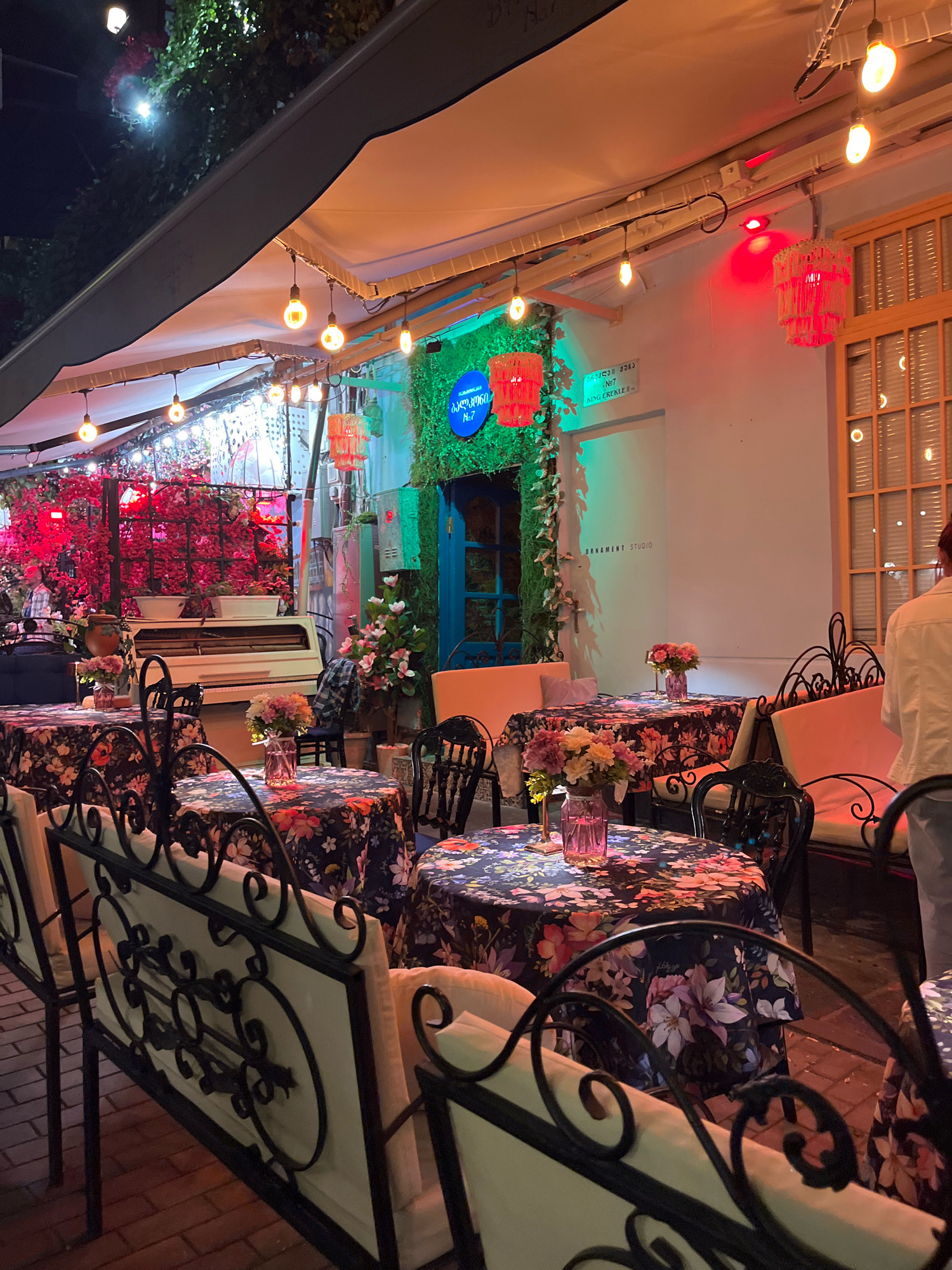 The image depicts an outdoor seating area of a restaurant or cafe in Tbilisi, Georgia, at night. The scene is illuminated by string lights and decorative lamps, creating a warm and inviting ambiance. Several tables are set with floral-patterned tablecloths and adorned with small vases of flowers. The furniture includes black metal chairs with ornate detailing and cushioned benches. In the background, a white piano is visible, adding a touch of elegance to the setting. The walls of the building are decorated with greenery and a blue door, suggesting a charming and possibly artistic establishment. The specific location within Tbilisi is indicated by a sign on the wall that reads "M. Dvali str. Nº7" and "SH. EREKLE II", suggesting a location on Erekle II Street, a well-known street in the Old Town area of Tbilisi, known for its restaurants, cafes, and shops. A person, partially visible, is standing near a window.