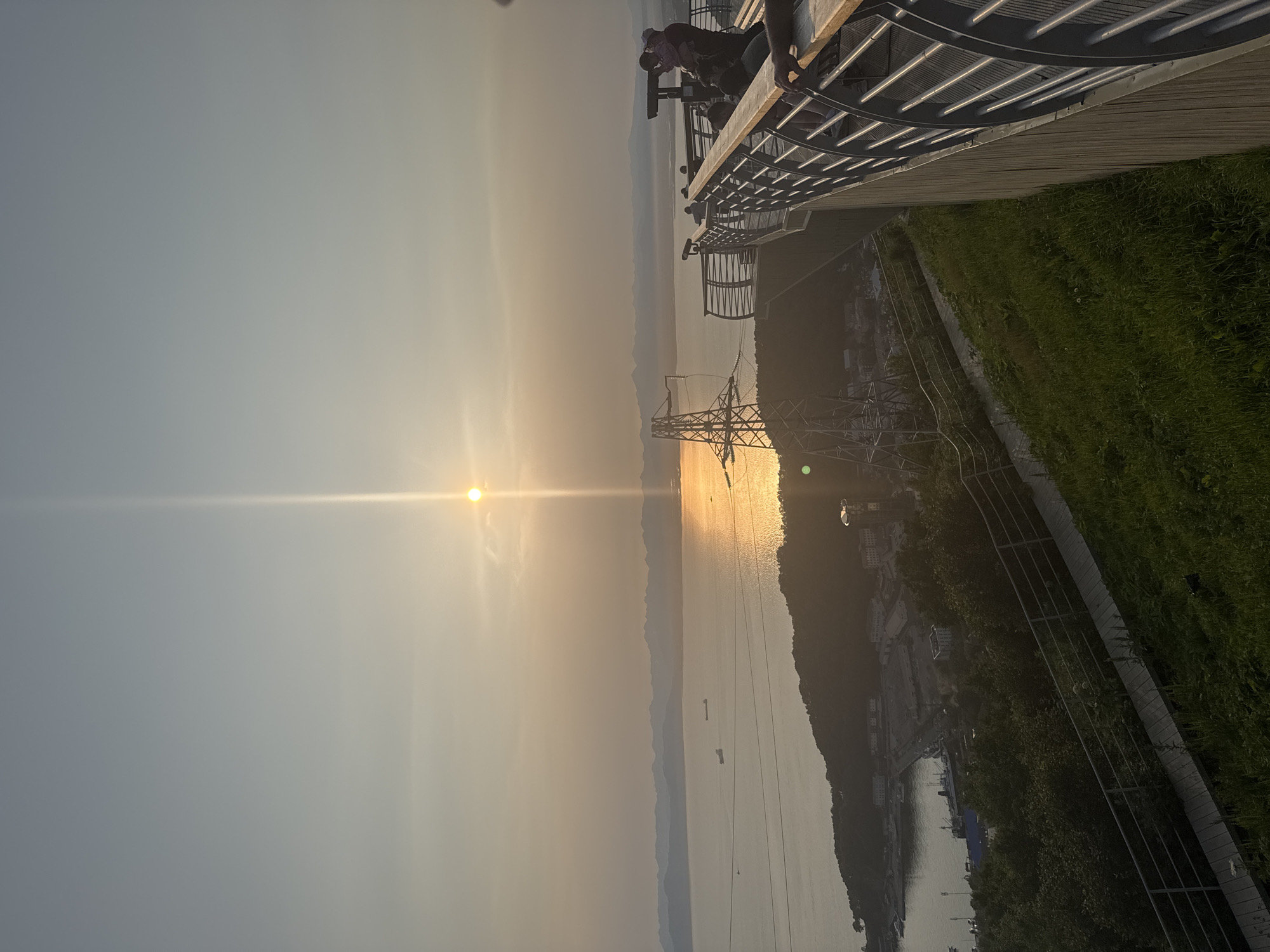 The image depicts a scenic overlook at sunset, likely in Petropavlovsk-Kamchatsky, Russia, offering a panoramic view of the Avacha Bay. In the foreground, a wooden platform with metal railings extends into the frame, serving as a vantage point for visitors. Several individuals are visible on the platform, some standing and observing the scenery, while one person is holding a child. In the middle ground, a steep, grassy hillside slopes down towards a bay area. The bay itself is dotted with several ships and boats, with buildings and infrastructure visible along its shoreline. Across the bay, distant mountains form a hazy silhouette against the sunset sky. A prominent latticed power transmission tower stands in the middle ground, its structure silhouetted against the bright sky. The sun is low in the sky, casting a warm, golden glow over the water, creating a reflective surface. The sky is a pale, hazy blue, with some wispy clouds. The overall impression is one of a peaceful, natural landscape viewed from a developed observation point.