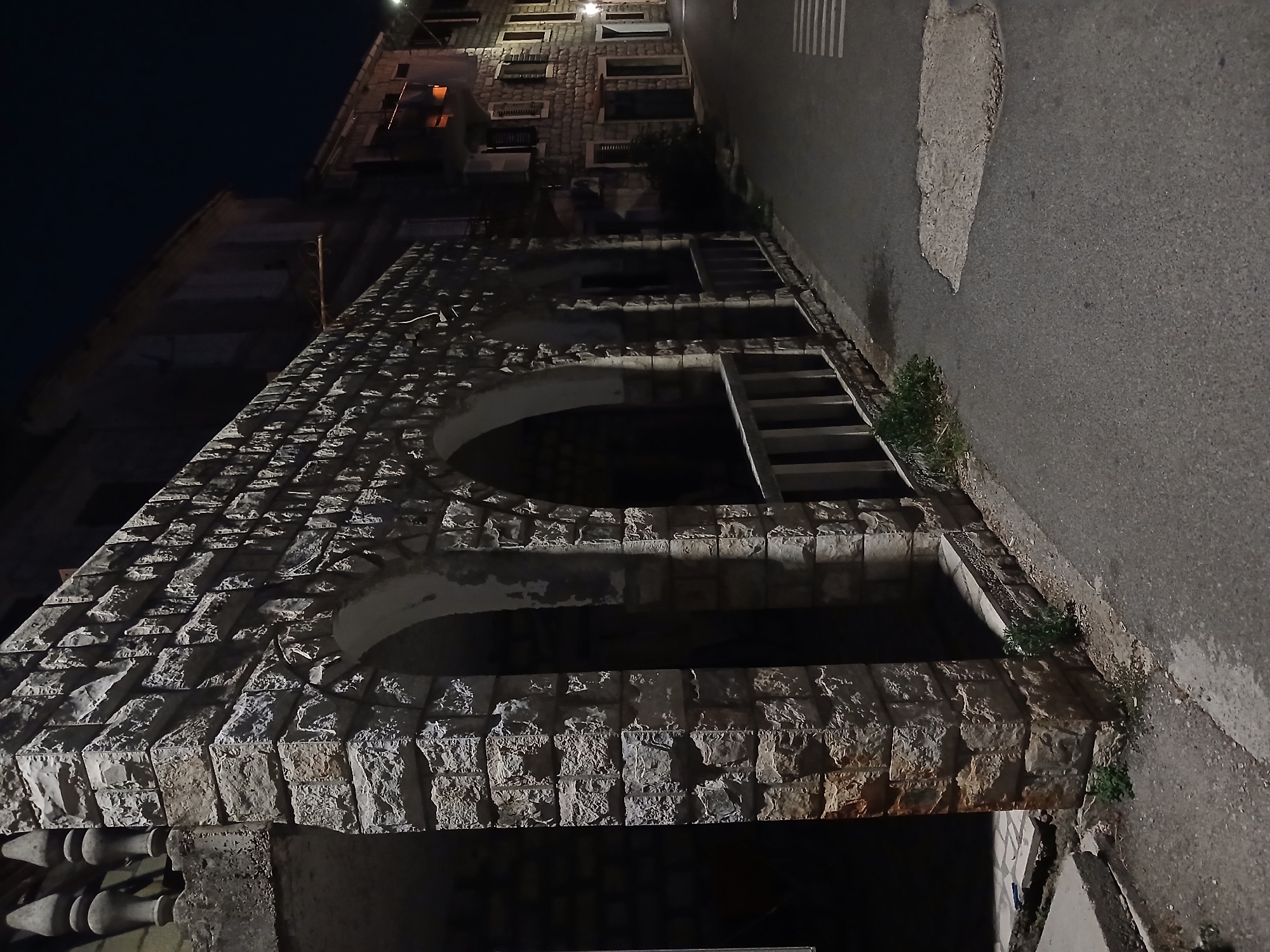 A night-time, eye-level, medium shot shows an old stone building with arched openings, situated on the left side of a paved street in an urban environment. The building's exterior is constructed with rough-hewn, rectangular stone blocks, some of which show signs of wear and weathering. An open walkway with a stone balustrade is visible within the arches. To the right of the building, the paved street curves away, and another stone building with windows and balconies is visible further down the street. The street is illuminated by artificial lights, casting shadows and highlighting the textures of the stone architecture. The sky is dark, indicating nighttime. The scene appears to be a quiet street in a historical European town.