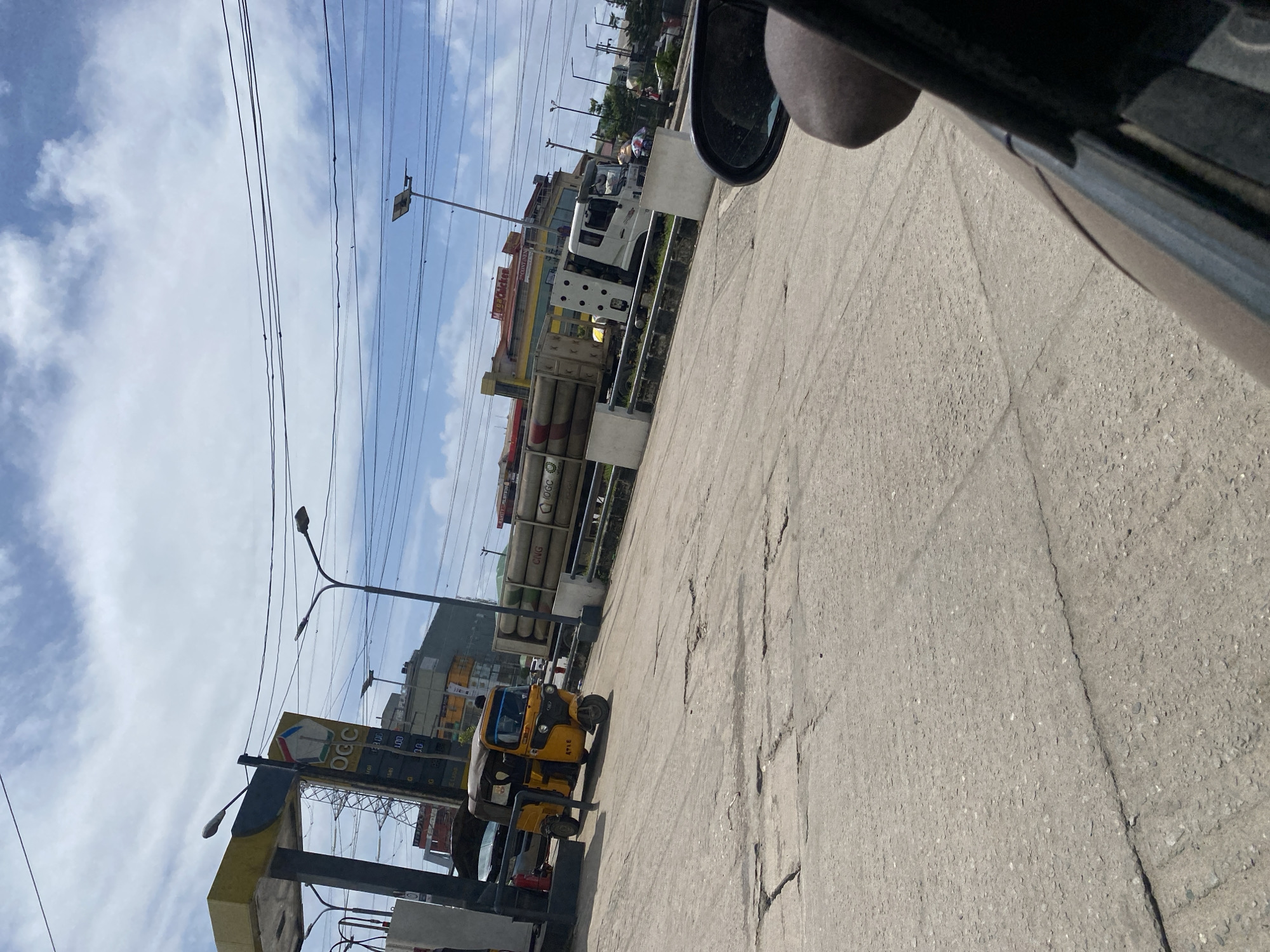 The image depicts a scene at a gas station in Ajah, Nigeria. In the foreground, a cracked concrete road is visible, with the side-view mirror and part of a car's interior on the right side of the frame suggesting the photo was taken from a moving vehicle. A yellow auto-rickshaw is parked at the gas station, its front facing left. Behind the auto-rickshaw, a white truck is parked, carrying a load of long, cylindrical tanks, some of which are labeled "CNG" and "IOGC". A gas station canopy with a price board displaying "OCC" and fuel prices is also present. In the background, several buildings, including one with "Meat & Chicken" signage and another with "CONTINENTAL" visible, are seen. Utility poles with wires crisscross the sky, which is partly cloudy.