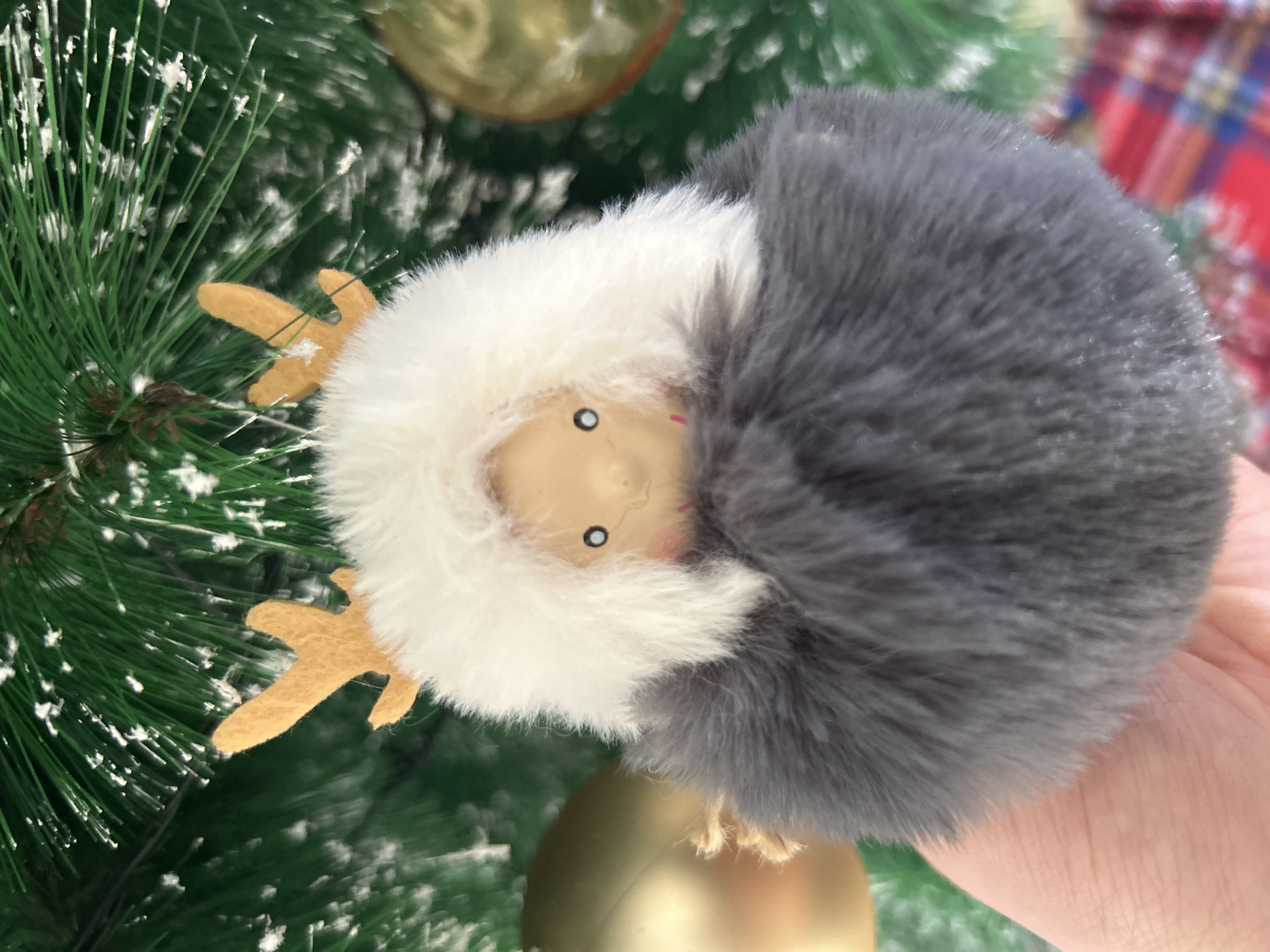 The image shows a reindeer Christmas ornament on a tree. The ornament has a small, tan-colored face with simple black eyes and a pink blush on the cheeks. It wears a white fluffy hood, partially covered by a larger, gray fluffy hood. The ornament has felt antlers that are light brown. The ornament hangs on a green Christmas tree with fake snow on the needles. In the background, there are gold ornaments and a glimpse of a plaid object.
There is no indication the image was taken in Câmara de Lobos, Portugal.