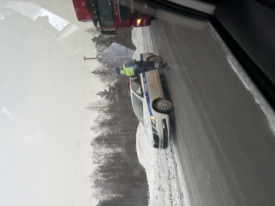 The image depicts a winter roadside scene in Yus'ki, Russia. A white and blue Russian police Skoda sedan, marked with "ПОЛИЦИЯ" (POLITSIA) on its side and displaying active red and blue emergency lights, is stationary on the right side of a snow-dusted asphalt road. The vehicle's license plate, "M276TA 777RUS," is visible. A male police officer stands near the driver's side of the police car, wearing a dark uniform, a dark winter hat, and a highly visible neon yellow-green reflective vest. His left arm is slightly bent, and his right arm is mostly obscured. To the immediate right of the police vehicle, the front right portion of a red Sitrak semi-truck with illuminated headlights is visible, partially obscured by the interior frame of the vehicle from which the photograph was taken. The environment features snow-covered ground, forming banks along the road, and a dense forest of bare deciduous and evergreen trees in the background under an overcast grey sky. Power lines and a utility pole are also visible in the middle ground. The road surface appears wet or slushy, indicating cold conditions.
