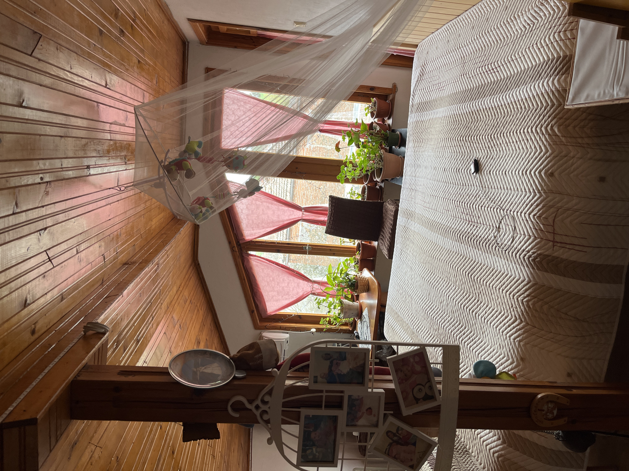 The image shows the interior of a bedroom in a likely rural setting, possibly in Polski Izvor, Bulgaria, given the prompt's instruction. The room has wooden walls and ceiling, giving it a rustic feel. A bed with a white patterned cover occupies a significant portion of the space. Windows with red valances are partially visible, allowing some natural light into the room, and several potted plants are situated near the windows. A white decorative frame with multiple picture frames is in the foreground. A clock is visible on a wooden beam. The scene appears domestic and lived-in.