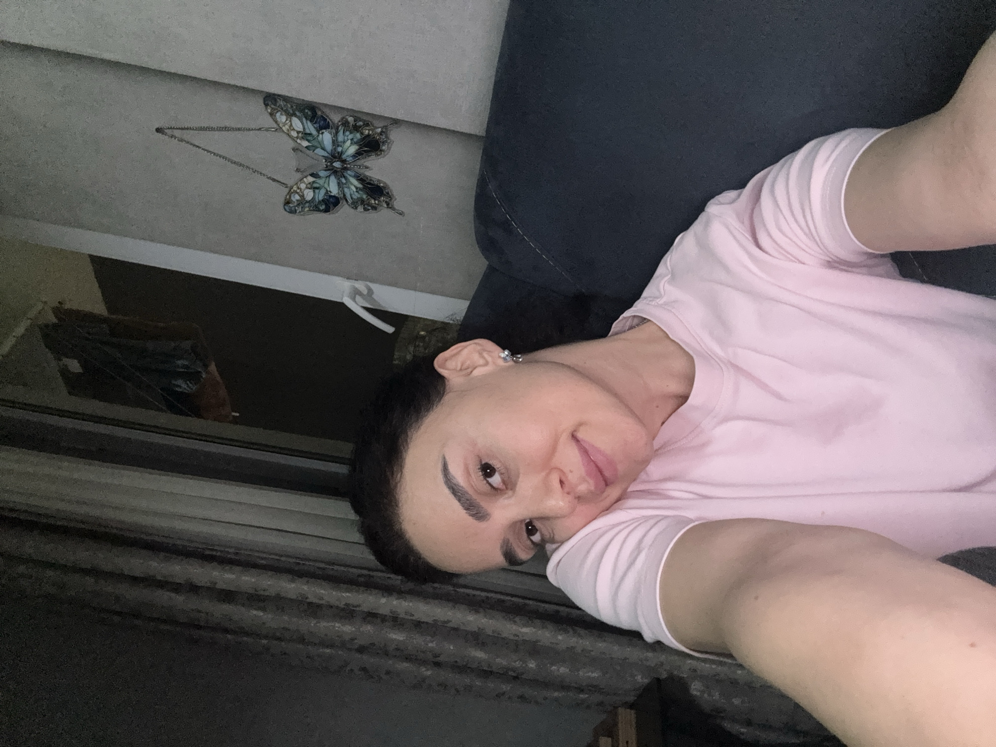 A woman is photographed indoors. She has dark hair, light skin, and full eyebrows. She is wearing a light pink t-shirt and an earring is visible in her left ear. She appears to be resting on a dark grey or black sofa. Behind her, hanging on a light grey or white wall, is a decorative butterfly piece. A window with a curtain is partially visible in the background. The location is within an interior, but there are no clues in the image to pinpoint it specifically within Tbilisi, Georgia.