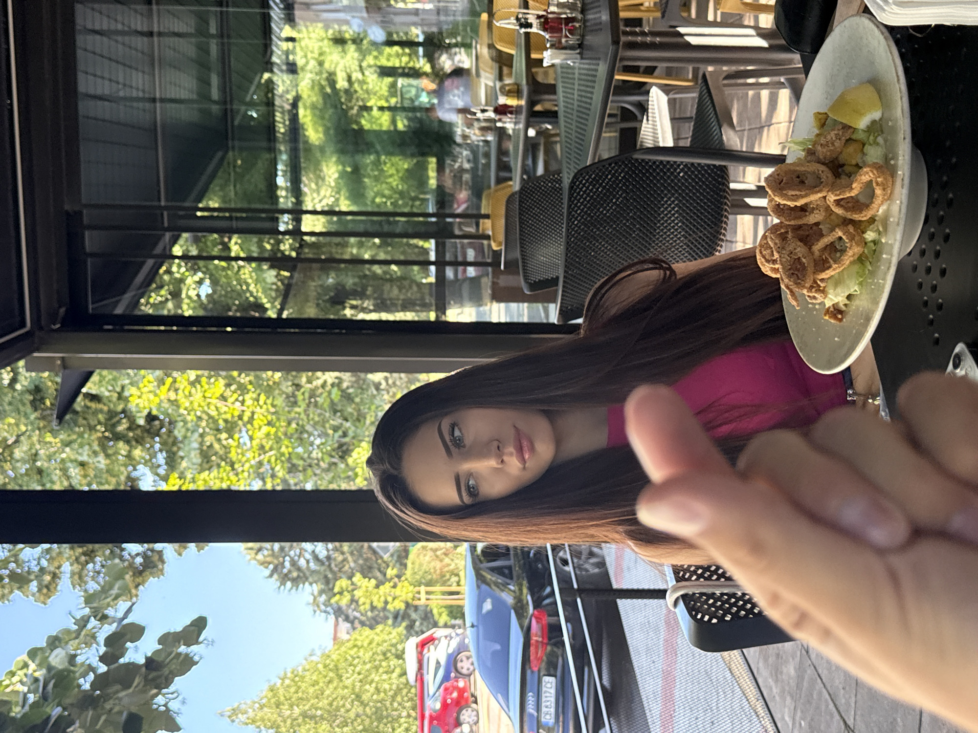 A young woman with long, dark hair and striking blue eyes is seated at an outdoor table at a restaurant. She is wearing a bright pink top. In the foreground, a hand is making a heart shape with its fingers. In front of the woman is a plate of fried calamari served on a bed of lettuce with a lemon wedge. The background shows parked cars, trees, and other tables and chairs at the restaurant, suggesting an urban setting. The overall scene depicts a casual meal on a sunny day.