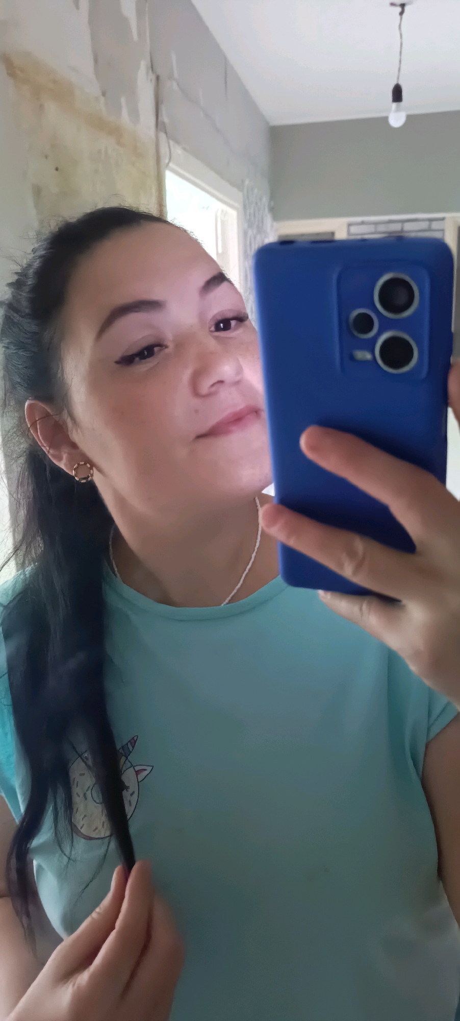 A woman is taking a selfie in a room. She has dark hair pulled back in a ponytail and is wearing a light blue t-shirt with a unicorn print. She is also wearing a white beaded necklace and a gold hoop earring in her left ear. She is holding a blue smartphone in her right hand, pointed towards herself. The background shows a wall that appears to be under renovation, with some peeling paint and exposed plaster. There is also a window and a lightbulb hanging from the ceiling. The room appears to be an interior space, possibly a residential apartment in Chaikovsky, Russia.