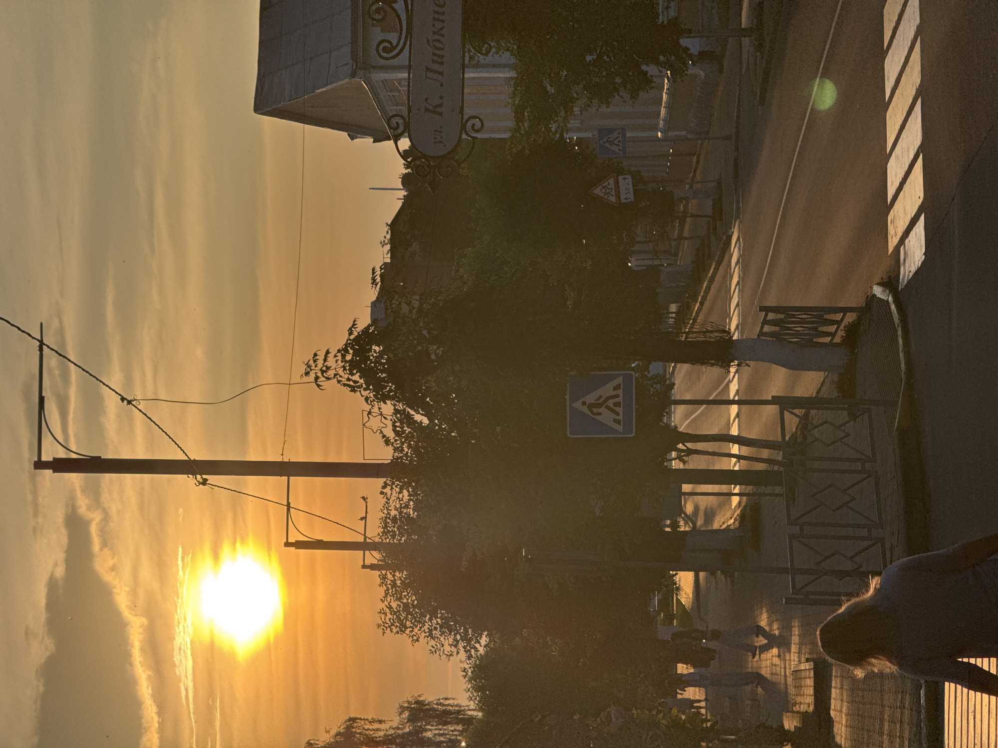 The image depicts a street scene in Nyasvizh, Belarus, at sunset. The sun, a bright orb, is low on the horizon, casting long shadows across the street and illuminating the sky with warm, orange hues. The street is paved, with a pedestrian crossing visible in the foreground and middle ground. A sidewalk runs along the left side of the street, lined with trees and a metal fence. Several people are walking on the sidewalk. On the right side of the street, buildings are visible, one of which has a sign that reads "ул. К. Любкне". Streetlights and utility poles are present along the street. The overall atmosphere is tranquil and evocative of a late summer evening.