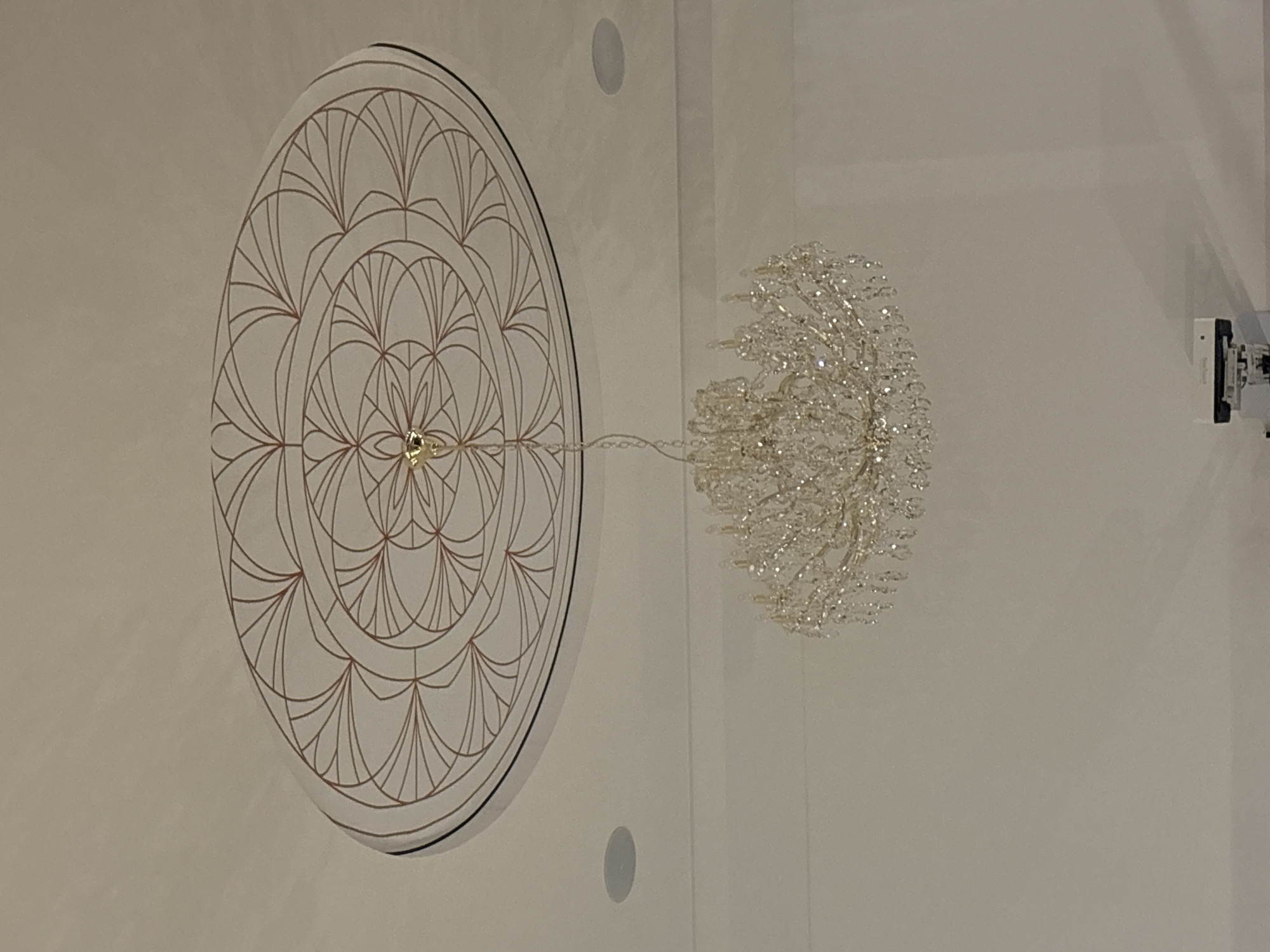 The image shows a ceiling with a decorative circular pattern and a crystal chandelier. The circular pattern, possibly a ceiling medallion, has a geometric design in a light brown color on a white background. The crystal chandelier, with its many sparkling crystals, is suspended from the center of the medallion. The background is a white ceiling with two small circular objects. The presence of a camera or sensor device, visible on the right, indicates the location might be under surveillance. Without additional information, I cannot confirm that this image was taken in Minsk, Belarus.