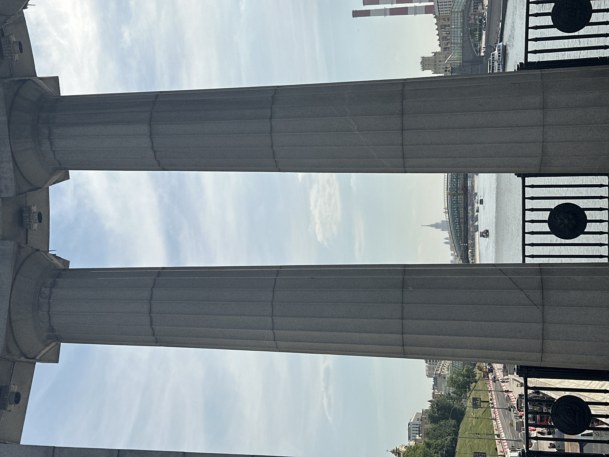 The image displays an urban riverine landscape in Moscow, Russia, viewed between two prominent, fluted, gray stone columns. These classical-style columns, detailed with capitals and horizontal shaft banding, form part of a balustrade or architectural overlook in the foreground.
Between the columns, the Moskva River flows from left to right, occupied by several boats, including at least two multi-deck white passenger ferries. On the far side of the river, an elaborate bridge with green-painted metal arches and supporting structures, possibly incorporating a covered walkway, spans the water. To the right of the bridge, two tall industrial chimneys, marked with alternating red and white horizontal stripes, rise prominently, indicative of a power plant (Moscow Central Heating and Power Plant No. 1, CHPP-1). Behind the right column and bridge, a multi-story building with a crenelated or turreted roof is partially visible.
On the near side of the river, to the left, an elevated embankment or roadway is apparent, lined with trees and streetlights. This area appears to be under construction or renovation, marked by orange barriers, construction equipment (including a small red excavator), and visible pedestrians. In the distance, above the left embankment, a cluster of buildings includes one with a small golden dome. Further downriver and centrally positioned on the distant skyline, a tall, multi-tiered Stalinist skyscraper with a prominent spire is visible (likely the Kotelnicheskaya Embankment Building). The sky above is a light blue with scattered wispy clouds. The scene is illuminated by natural daylight, suggesting late morning or afternoon.