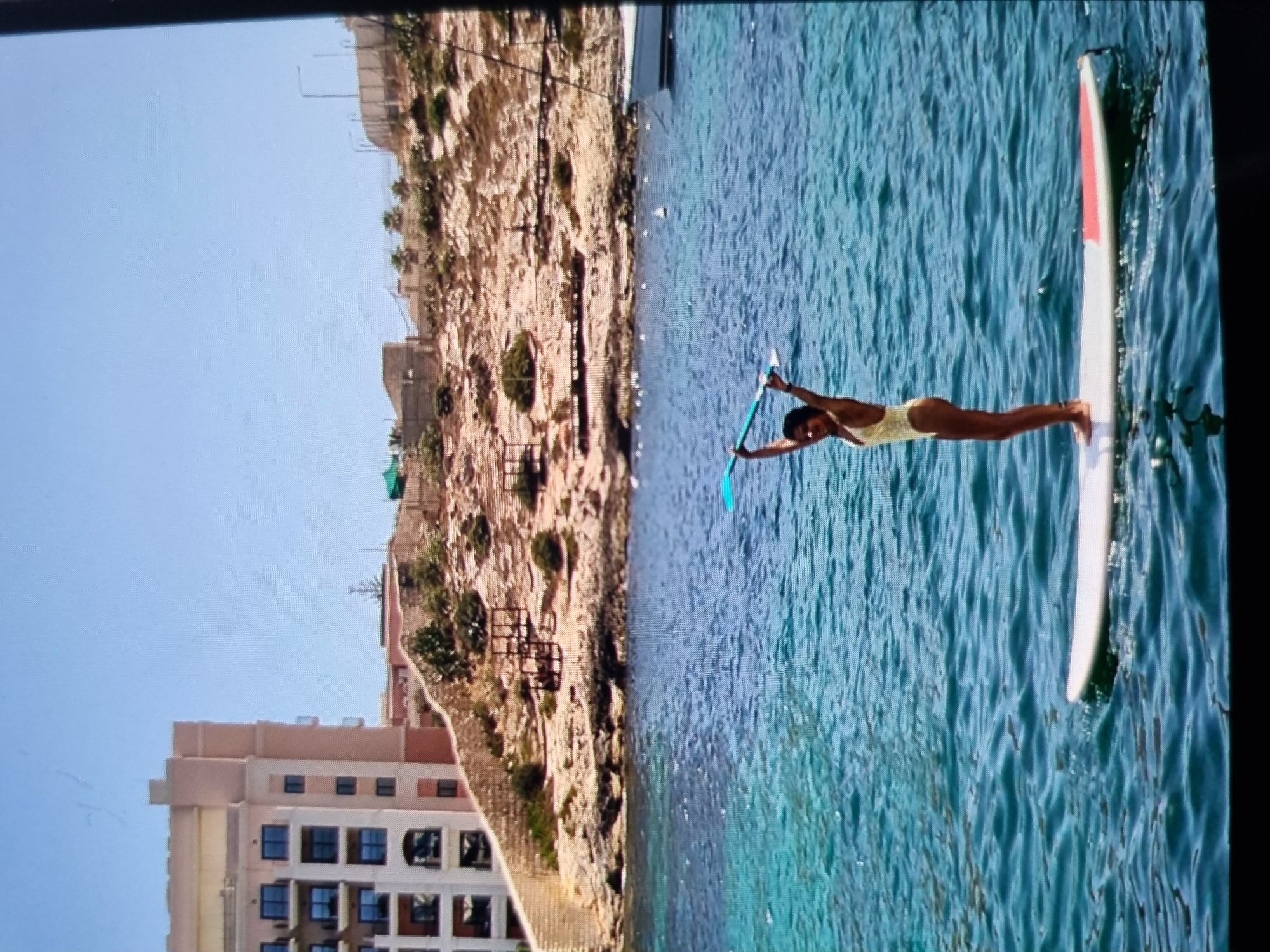 A woman stands on a paddleboard in turquoise water near a rocky coastline. She is using a paddle to move the board. A building is visible on the coastline. There is no evidence that this photo was taken in Plovdiv, Bulgaria.