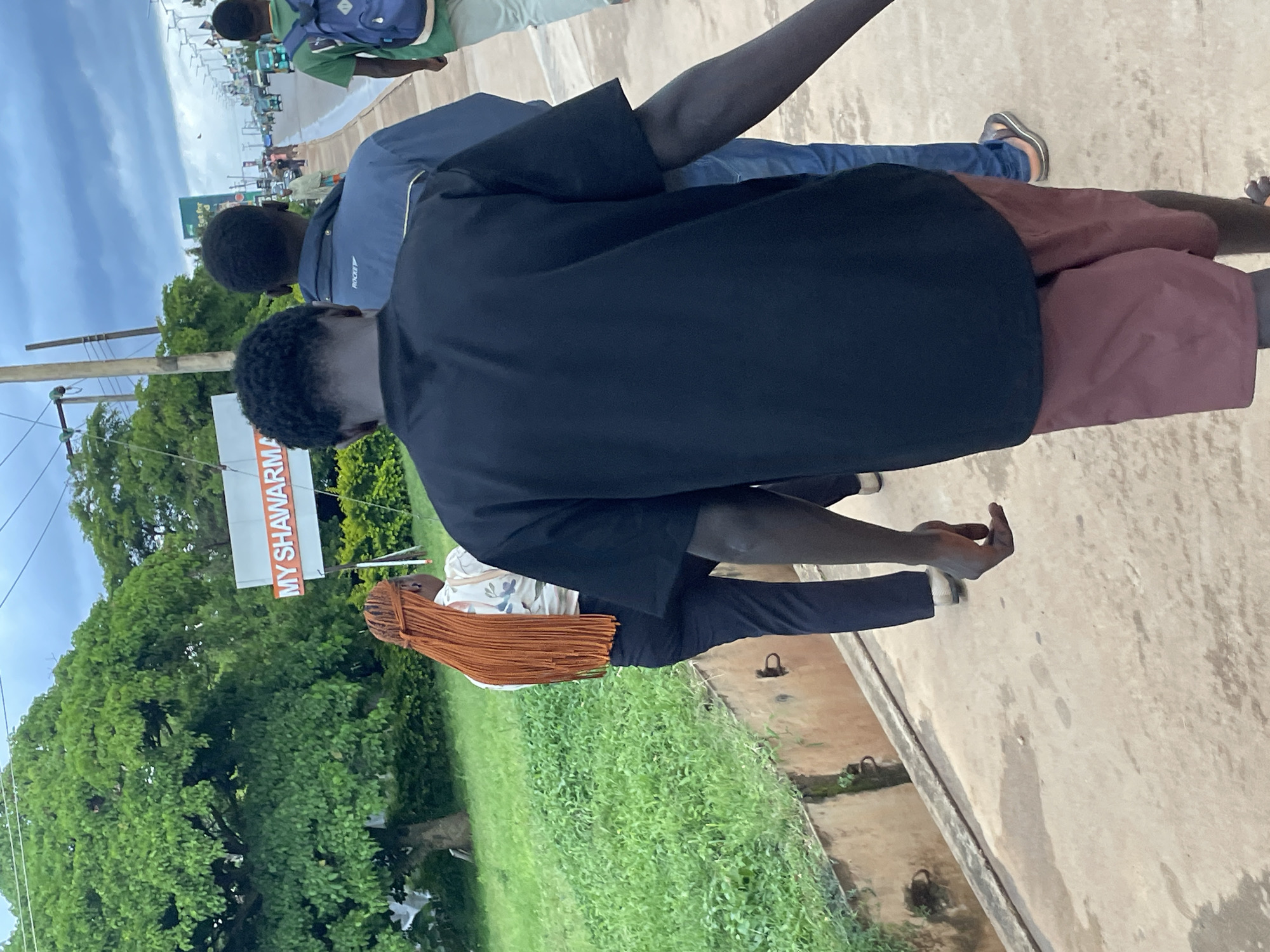 In the city of Jos, Nigeria, a group of individuals walks along a paved sidewalk adjacent to a grassy embankment. In the foreground, a person wearing a black t-shirt and maroon shorts is observed from behind, their right arm extended slightly. To their left, a person with long, auburn braided hair wears a patterned shirt and dark trousers. Further ahead, two men are visible; one in a blue jacket and jeans, and another in a green t-shirt carrying a blue backpack. Behind them, the sidewalk continues, lined with streetlights and a busy road with various vehicles, including motorcycles and cars. A sign displaying "MY SHAWARMA" is visible among the trees and foliage. The sky is overcast with clouds, suggesting a potentially humid or rainy day.