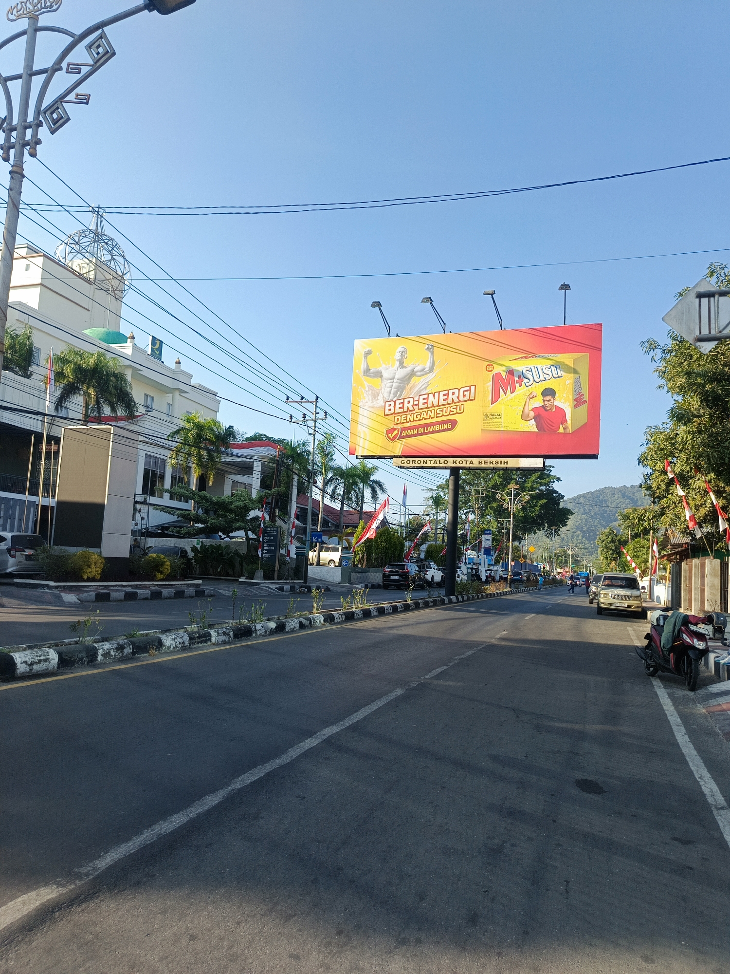 The image captures a bright, sunny day in Gorontalo City, Indonesia, with a clear blue sky. A wide road dominates the foreground, bordered by a raised median strip with sparse vegetation and a black and white striped curb. On the left side of the road, a modern, multi-story building with a green dome is visible. Palm trees line the front of the building, and Indonesian flags are displayed along its facade.
A prominent billboard stands on the right side of the road, advertising a milk product named "M Susu". The billboard features images of a muscular man and a man in a red shirt, with text in Indonesian promoting energy and stomach health. Below the billboard, the slogan "GORONTALO KOTA BERSIH" (Gorontalo Clean City) is displayed.
The road is moderately busy with various vehicles, including cars and a motorcycle parked on the right side. In the background, lush green hills and mountains are visible, adding to the scenic beauty of the location. Electrical wires crisscross the sky, a common sight in urban Indonesian landscapes. The overall impression is that of a vibrant and active city environment.