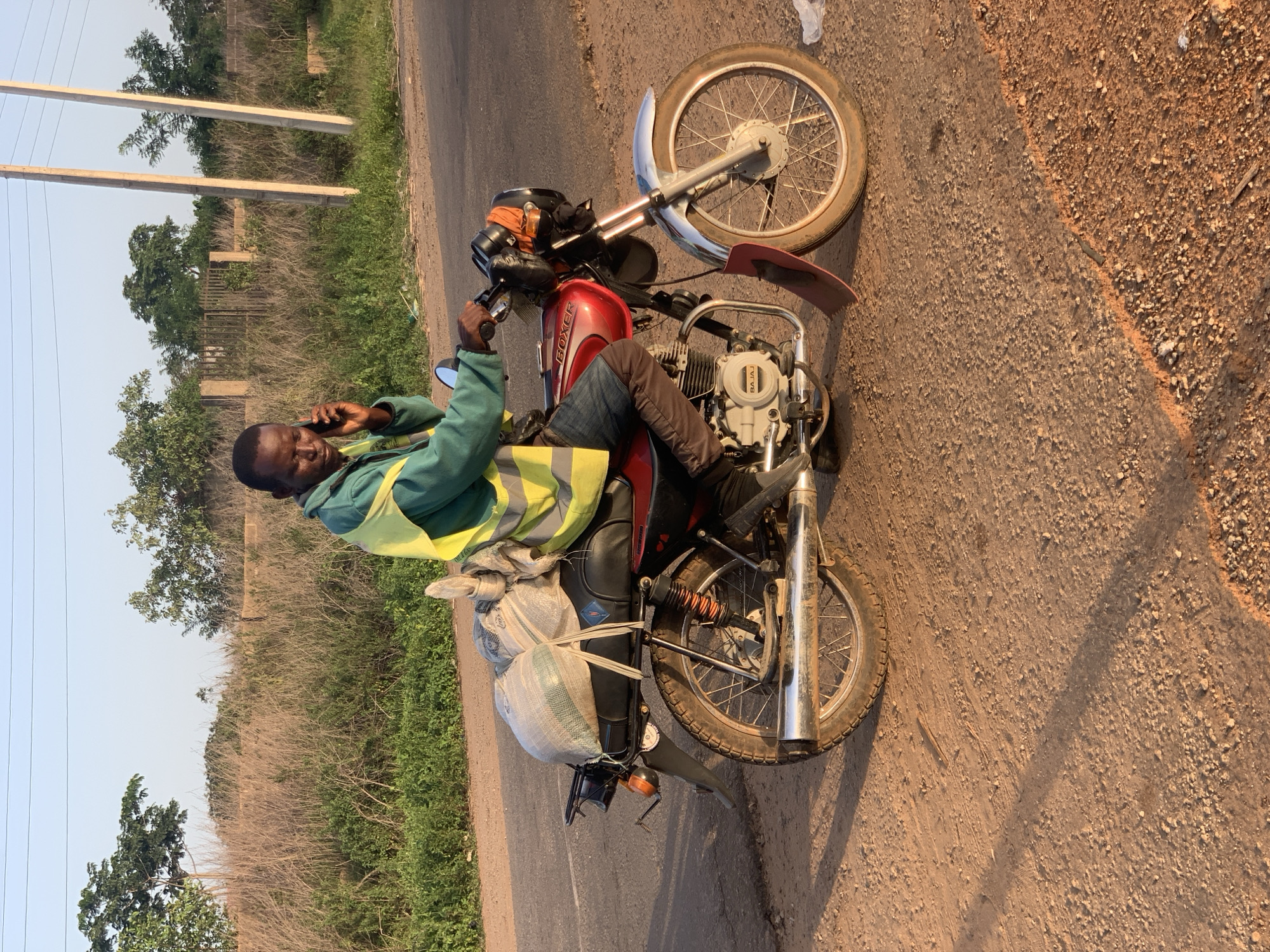 A man in a neon yellow safety vest is sitting on a red "BOXER" brand motorcycle on a paved road with a dirt shoulder. The motorcycle is carrying a large bundle of bagged items tied to the back. The man is holding a cell phone to his ear. The background features tall grass, trees, and utility poles under a clear sky. The image is likely taken in Kabba, Nigeria.