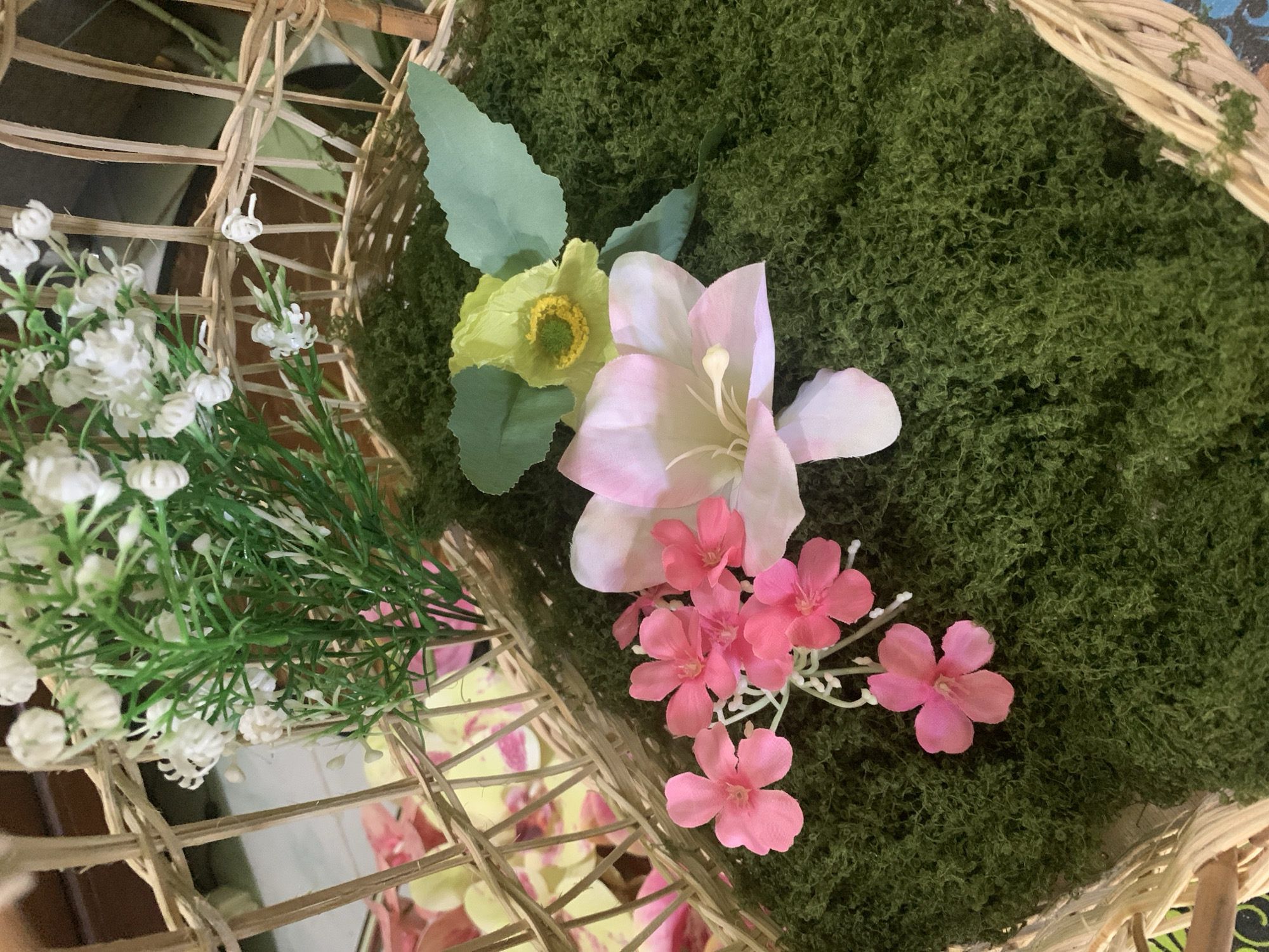 The image displays an arrangement of artificial flowers and greenery within a wicker basket. The basket is partially filled with a textured, dark green material that resembles moss. Several artificial flowers are placed on top of the moss. To the left, a cluster of small, bright pink flowers with five petals each are arranged on delicate stems. In the center, a larger, pale pink and white flower with prominent stamens is visible. To the right, a light yellow flower with green accents and two large, light green leaves are positioned. In the background, behind the flowers and moss, a bunch of small white artificial flowers with green stems emerges from the top of the basket. The wicker basket itself is a natural, light brown color and appears to be woven with thin strands. In the blurred background, there are hints of other floral decorations, including what appears to be pale pink and yellow artificial flowers peeking through the wicker. The scene suggests a decorative arrangement, possibly for a special occasion or as a home decor item. The image lacks any context or specific details that would place it in Kuala Pembuang, Indonesia.