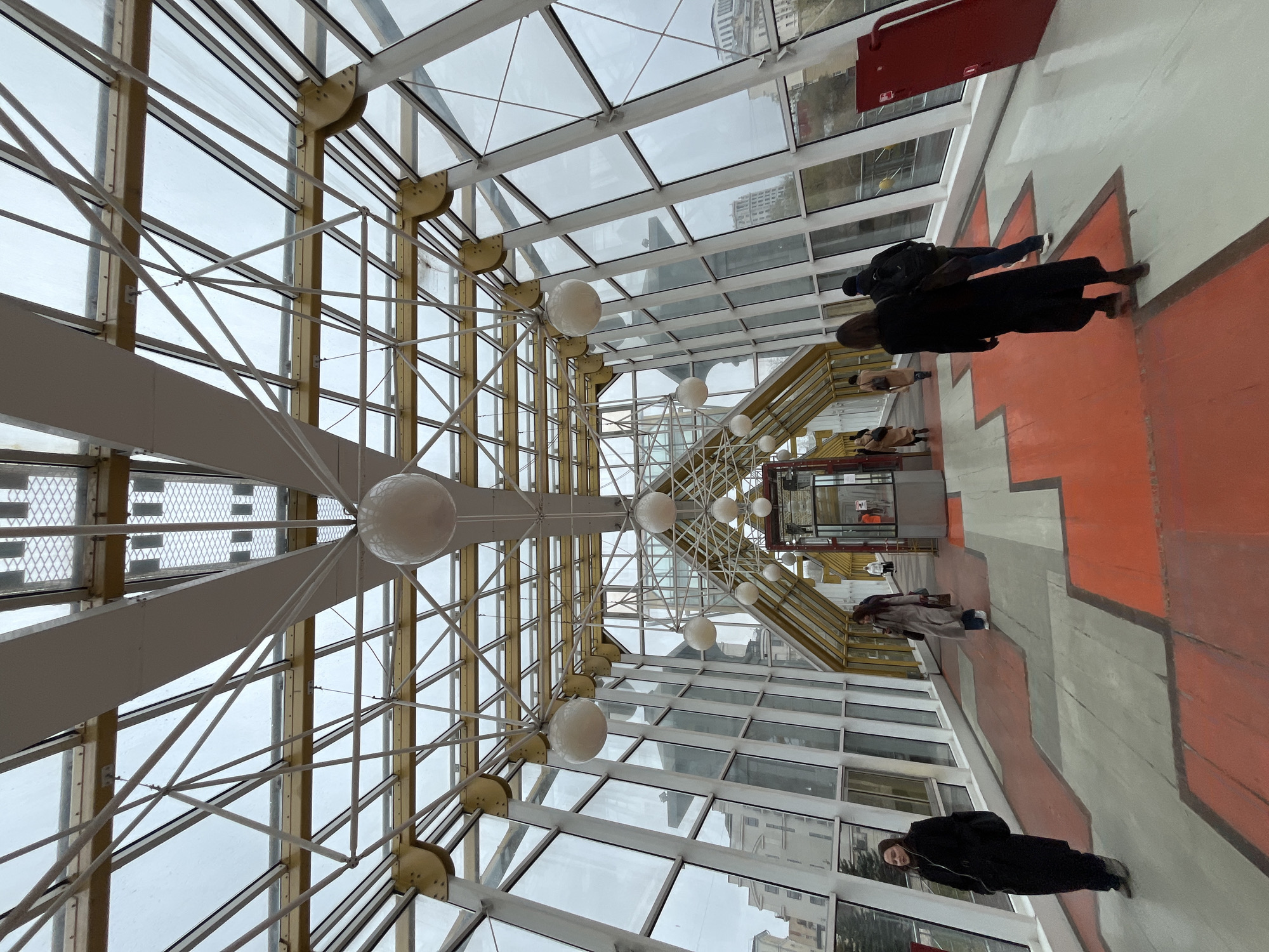 The image shows the interior of a glass-roofed walkway or corridor in Moscow, Russia. The architectural style is characterized by a modern, industrial aesthetic, featuring a complex network of white and gold-colored metal beams supporting the glass roof. Round, white light fixtures hang from the ceiling. The floor is patterned with red and gray geometric shapes. Several people are present, walking through the corridor. The image is taken from a low angle, looking upwards toward the ceiling and roof. The view through the glass roof reveals overcast skies and glimpses of buildings outside.