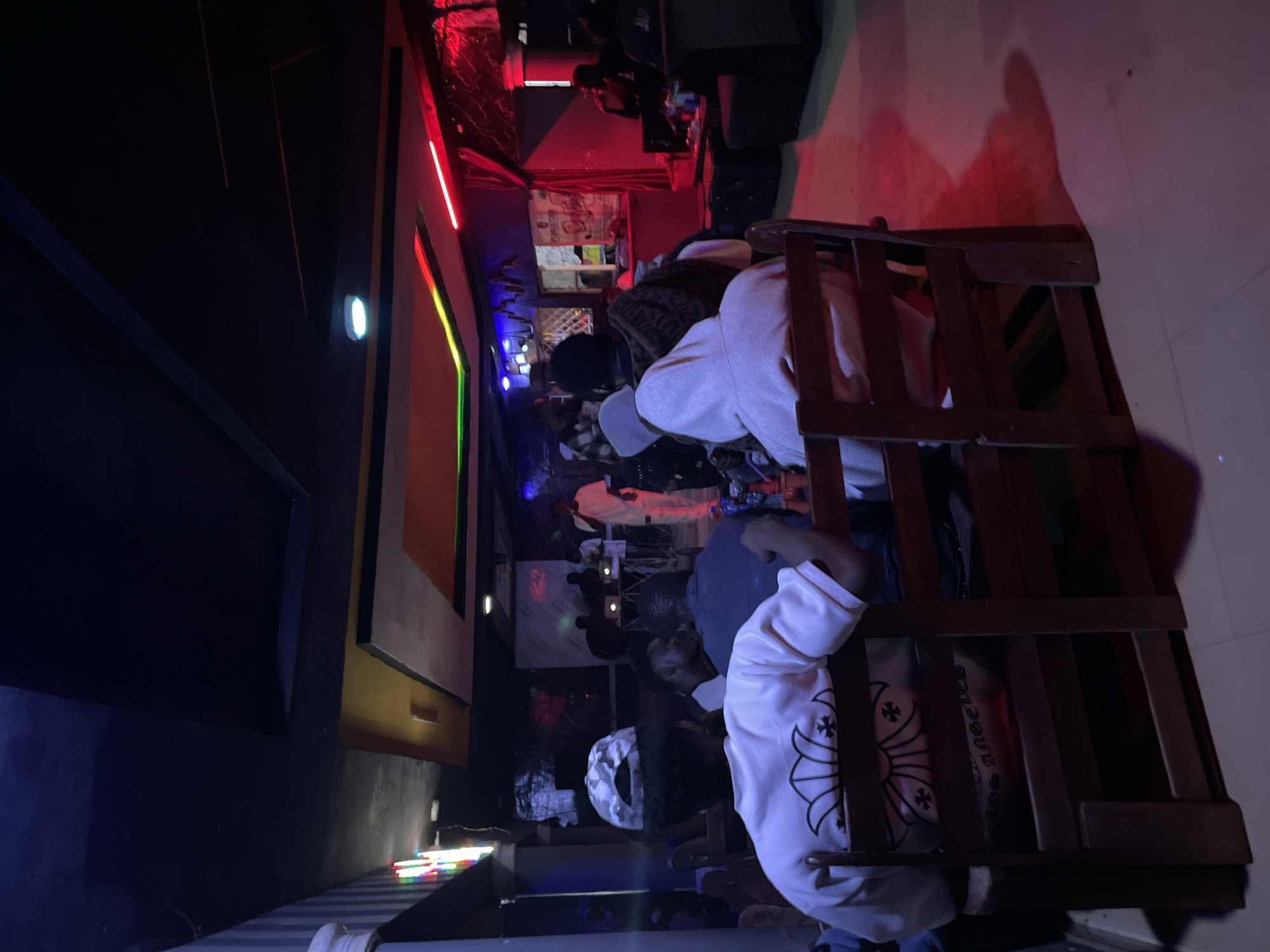 The image depicts an indoor scene, likely a lounge or entertainment venue, illuminated by a combination of ambient and colored lighting. In the foreground, two individuals are seated facing away from the camera, with their backs to a wooden chair. One person is wearing a white t-shirt with a graphic design and a camouflage baseball cap. The other person is wearing a light-colored hooded sweatshirt.
In the mid-ground, a group of people are gathered, some standing and others seated. A stage or performance area is visible further back, with individuals on it, possibly engaged in a musical performance or DJ set. There are speakers and equipment on the stage.
The environment appears to be a dimly lit establishment with a dark ceiling featuring recessed lighting and neon accents in red, green, and blue. The walls have a dark finish. The floor is tiled. The overall impression is that of a nighttime social gathering or event. The image does not provide any explicit information about the city of Durimi, Nigeria, or any specific location within it.