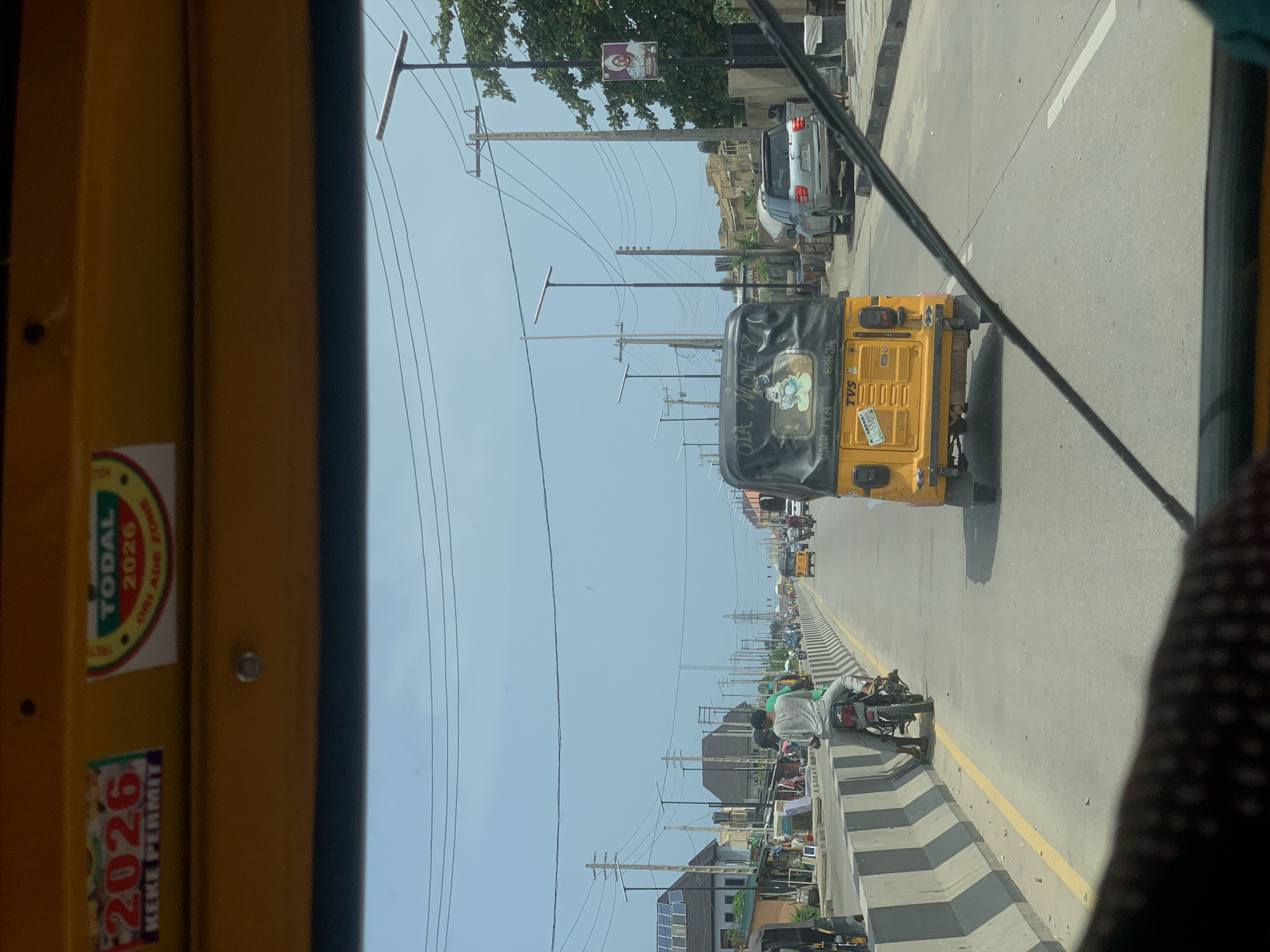 The image shows a view from inside a yellow vehicle, likely a "Keke Napep" (a type of auto-rickshaw), traveling along a road in Satellite Town, Nigeria. The "Keke" has stickers on the left interior, including one that says "2026 KEKE PERMIT" and another that says "TODAL 2026 ORE ADE ZONE". Another "Keke" is visible ahead, with writing in Yoruba "OLA MONEY, OMO IYA" and registration "MUS500PJ" clearly visible. On the right, there's a road divider with a motorcycle parked beside it, and a man in a white and grey striped shirt is working on the bike. Further down the road, there is traffic in both directions and streetlights along the road. There are also power poles with wires. The sky is clear. Buildings and solar panels are seen at the bottom of the image along the side of the road, likely a street market.