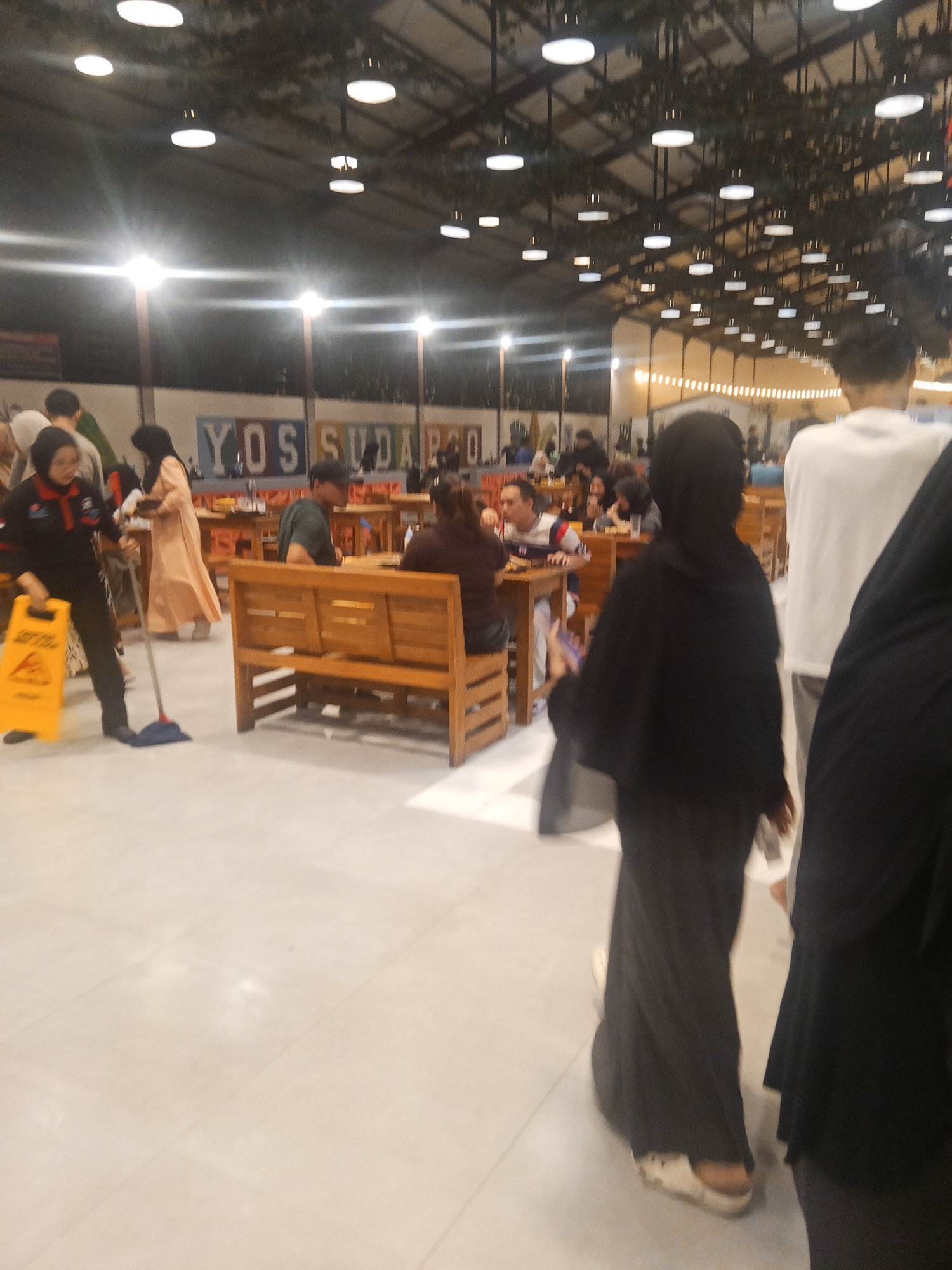 The image shows an indoor scene, likely a food court or restaurant, with several people visible. The location is identified as Lubuklinggau, Indonesia, specifically at "YOS SUDAKO" which appears to be the name of the establishment. In the foreground, a person in a black hijab and black abaya is walking, while another person in a white shirt and grey pants is walking away from the camera. To the left, a person wearing a black uniform with red accents is mopping the floor with a yellow wet floor sign nearby. Other individuals are seated at wooden tables and benches in the background, some eating and others conversing. The environment is well-lit with numerous round pendant lights on the ceiling, and decorative greenery hangs from the ceiling structure. The floor is tiled.