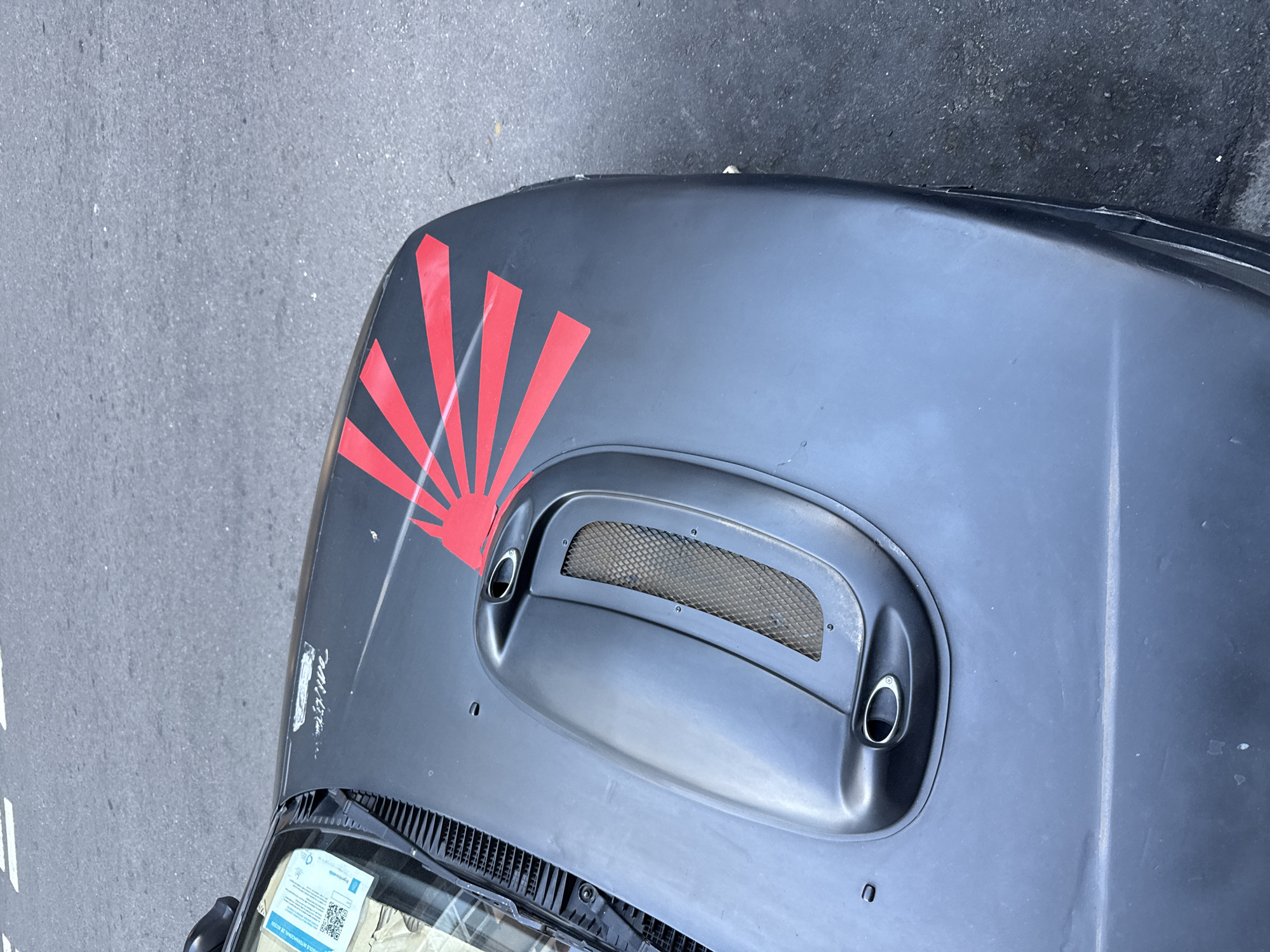 A high-angle shot shows the hood of a gray car parked on a street. The hood has a red design resembling rising sun rays on the left side and a raised vent in the center with a mesh screen. The vehicle is parked on a road with painted white lines. The hood also has various imperfections and scratches on the paint. A document with QR codes is visible on the dashboard. Based on the document visible on the dashboard, it is likely that the photograph was taken in Buenos Aires, Argentina.