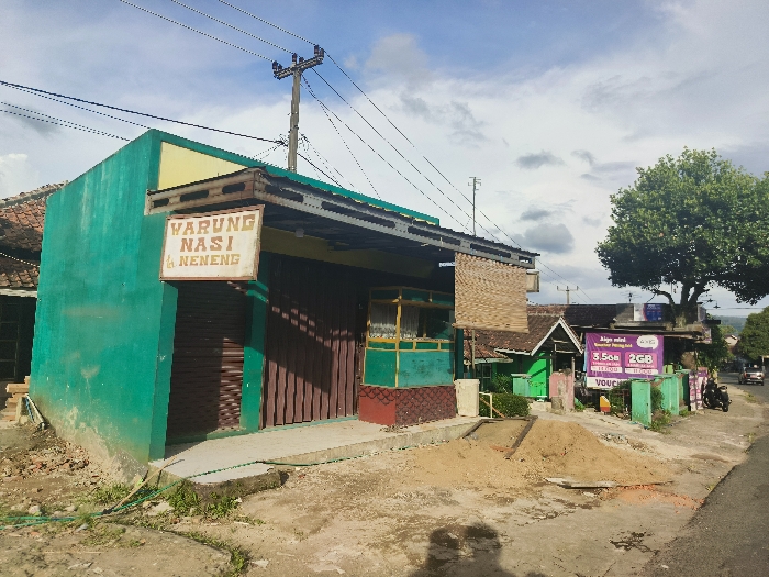 The image shows a small, one-story building with a green facade and a brown awning. The building has a sign that reads "Warung Nasi & Neneng." The building is located in Cikalong, Indonesia, at coordinates -6.758 latitude, 107.446 longitude. There is a dirt road in front of the building and a pile of dirt to the right of the building. A tree is visible in the background, to the right of the building. The street is paved. A sign for a telecommunications company is visible in the distance, along with a few vehicles. The building appears to be a small restaurant or shop. The scene is shot during the day under a clear sky.