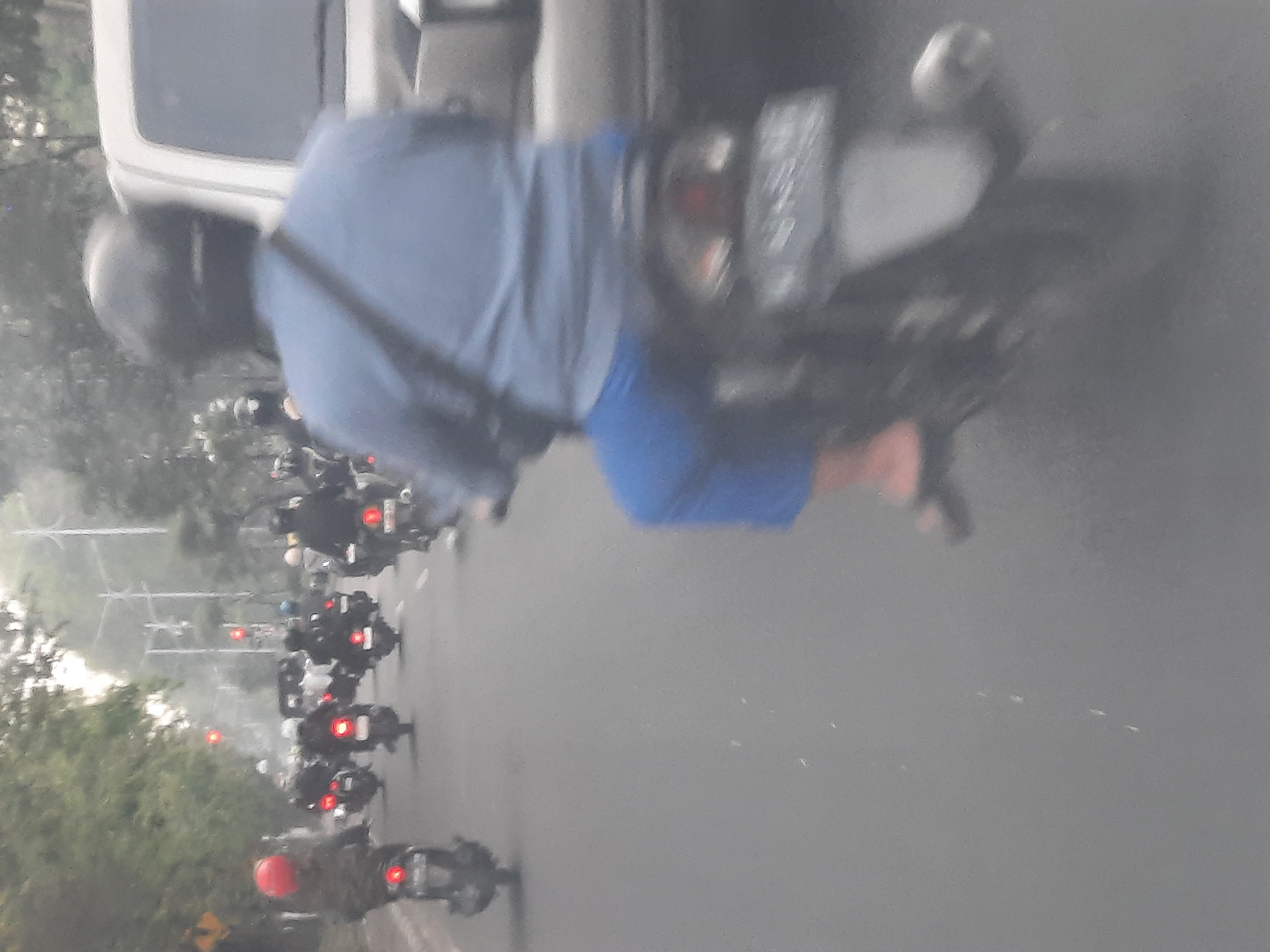 In Banjarkemantren, Indonesia, a dense traffic scene unfolds on a road. Multiple motorcycles are visible, carrying riders, many of whom are wearing helmets. One rider, in the foreground and slightly to the right, is wearing a blue shirt and blue pants and is on a motorcycle. To their left, another motorcyclist with a red helmet is seen. Other motorcycles with riders are scattered throughout the scene, some with their brake lights illuminated, indicating they are either stopped or slowing down. The background shows a row of trees and utility poles, suggesting an urban or suburban environment. The overall atmosphere appears to be a busy commute, possibly during daytime.