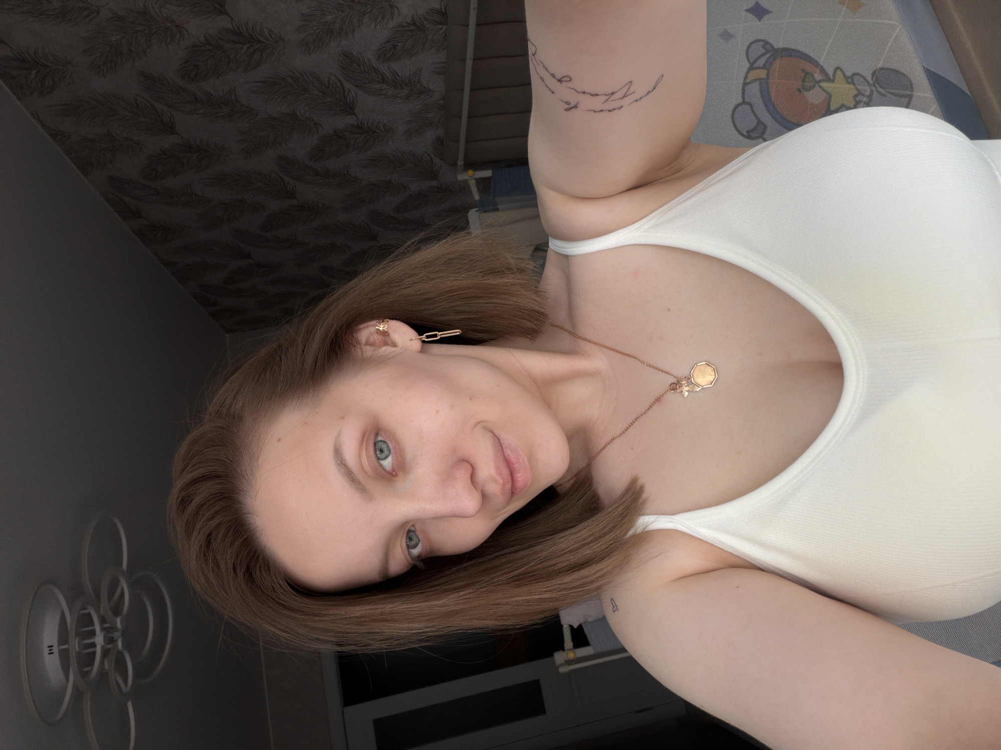 A selfie captures a young woman indoors in Reshetnikovo, Russia, positioned with her head tilted to the left, looking directly at the camera with a subtle, gentle smile. Her brown, medium-length hair is styled in a bob. She has blue-green eyes and fair skin. She wears a plain white tank top or camisole.
Her visible jewelry includes a delicate gold-colored chain necklace with two pendants: one is an octagonal shape, and the other appears to be a small bee or star charm. She also has multiple gold-colored earrings; one on her left ear is a small stud, possibly star-shaped, and another is a longer, paperclip-link style.
Several tattoos are visible on her left arm. On her upper bicep, script text reads, "do your own thing", or similar, partially obscured. A small outline of a heart is tattooed on her forearm, near the elbow.
The background suggests an indoor setting. Behind her head and shoulder, a dark wall features a repeating, subtle pattern of dark feathers or leaves. To her lower right, a light-colored mat or rug displays a cartoon character resembling a robot or astronaut with a star, in shades of orange, blue, and gray. On the far left, a modern-looking white light fixture composed of concentric rings is mounted on a dark wall. The overall lighting is even and bright, consistent with an indoor environment.