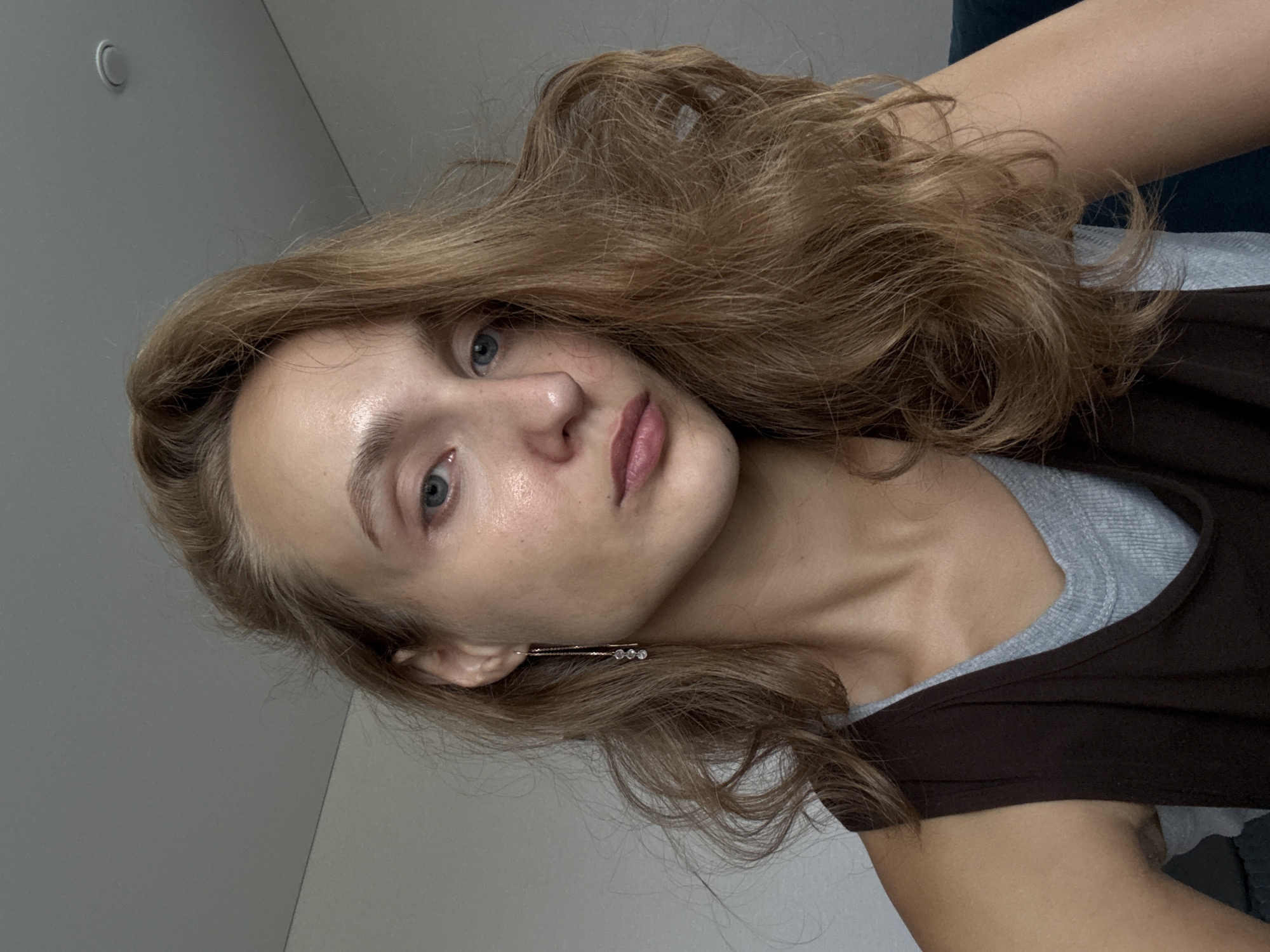 A close-up, waist-up, indoor portrait shows a young woman with light brown, wavy hair and blue eyes. She is looking directly at the camera with a neutral expression. She is wearing a dark brown tank top over a light grey ribbed t-shirt. A delicate gold earring with three clear beads hangs from her left ear. The background is a plain, light-colored wall. The lighting suggests an overcast day or indoor lighting. The woman appears to be in a room within a residential building. There is no discernible information about the location in the city of Подшивалово, Russia.