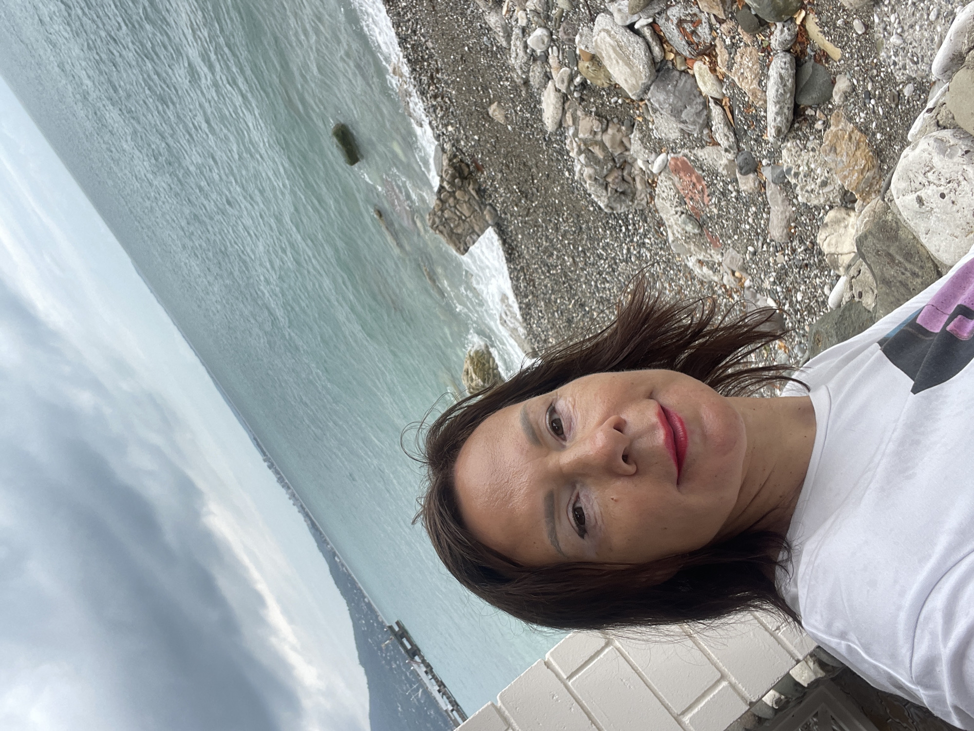 A woman with brown hair, wearing a white t-shirt, takes a selfie outdoors near a rocky beach. She is smiling and has red lipstick on. The background shows the turquoise sea under a cloudy sky. A distant coastline with hills and buildings is visible. Part of a white brick wall is in the foreground on the left. The beach is covered in grey pebbles and large rocks.
