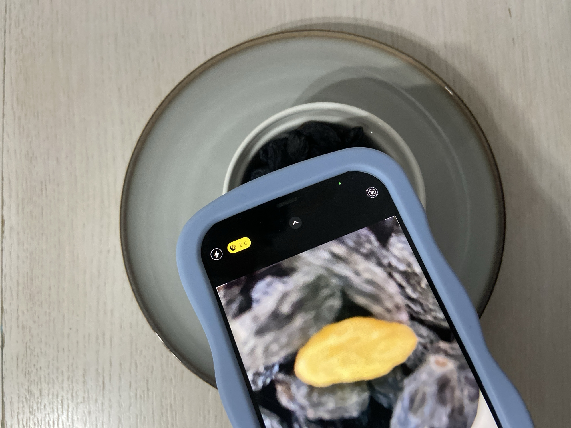 A mobile phone, encased in a blue case, is being used to photograph a dish. The dish rests on a white surface and contains a smaller bowl filled with what appears to be dark-colored food items. The phone screen displays a close-up view of what appears to be a yellow food item, possibly a dried fruit or candy. The scene is well-lit, and the focus is primarily on the phone and the dish. There is no indication of location, nor mention of Budva, Montenegro, in the source image.