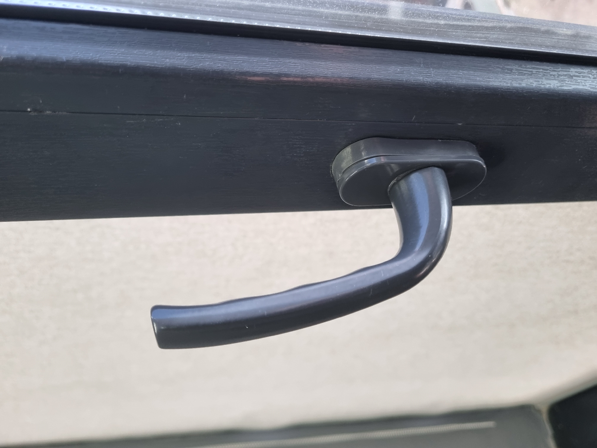A dark gray handle is attached to the frame of a dark gray window. The handle is curved and extends outwards from the frame. The background is a light-colored wall. The image is a close-up shot of the window handle. The location is not specified in the image.