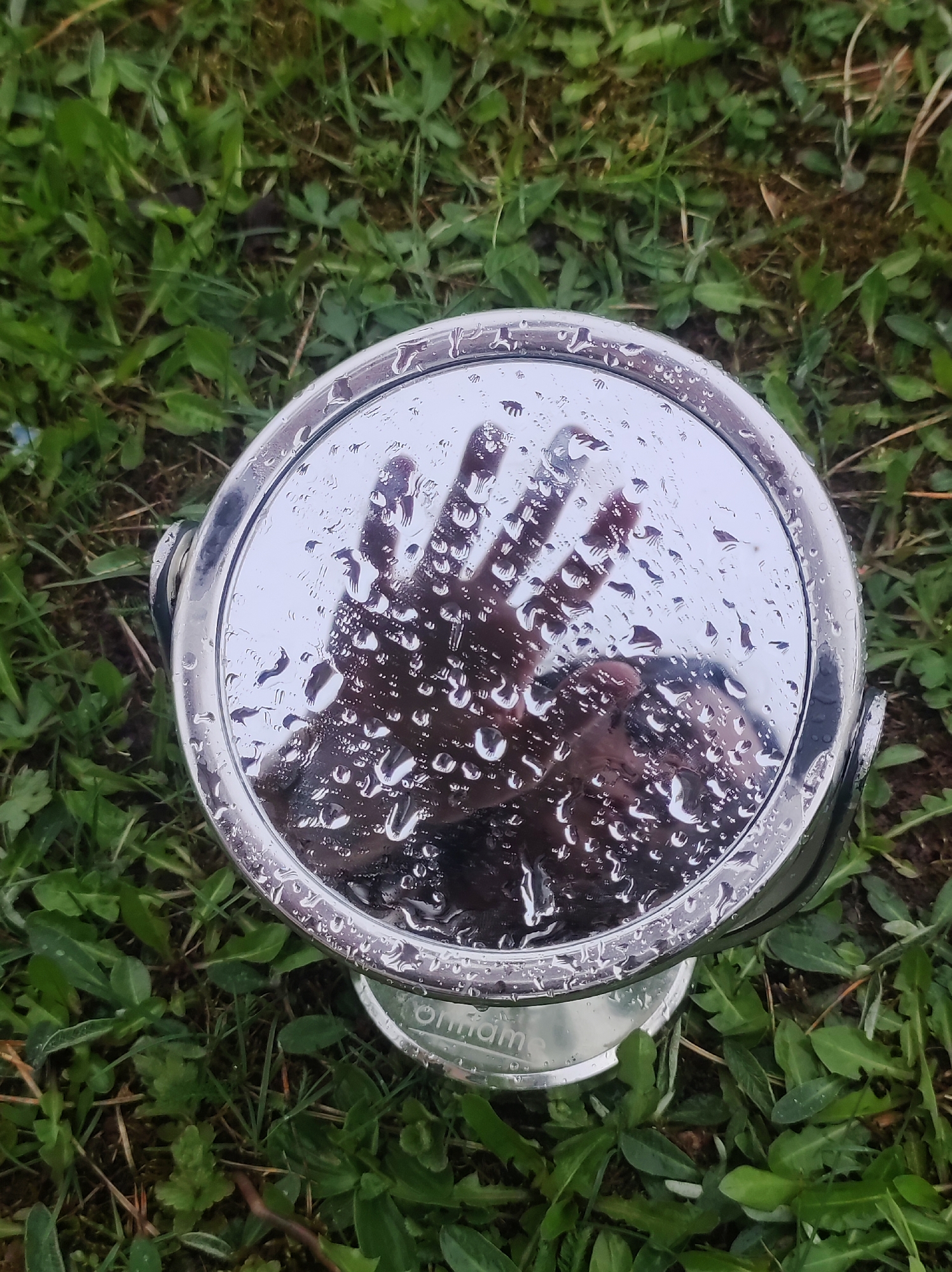 A circular, chrome-framed mirror, partially covered in water droplets, reflects a dark-skinned hand against a blurred background. The mirror rests on a stand, also chrome, sitting on a patch of green grass. The word "Onam" is visible on the stand. The location cannot be identified as Фоки, Russia from the image alone.