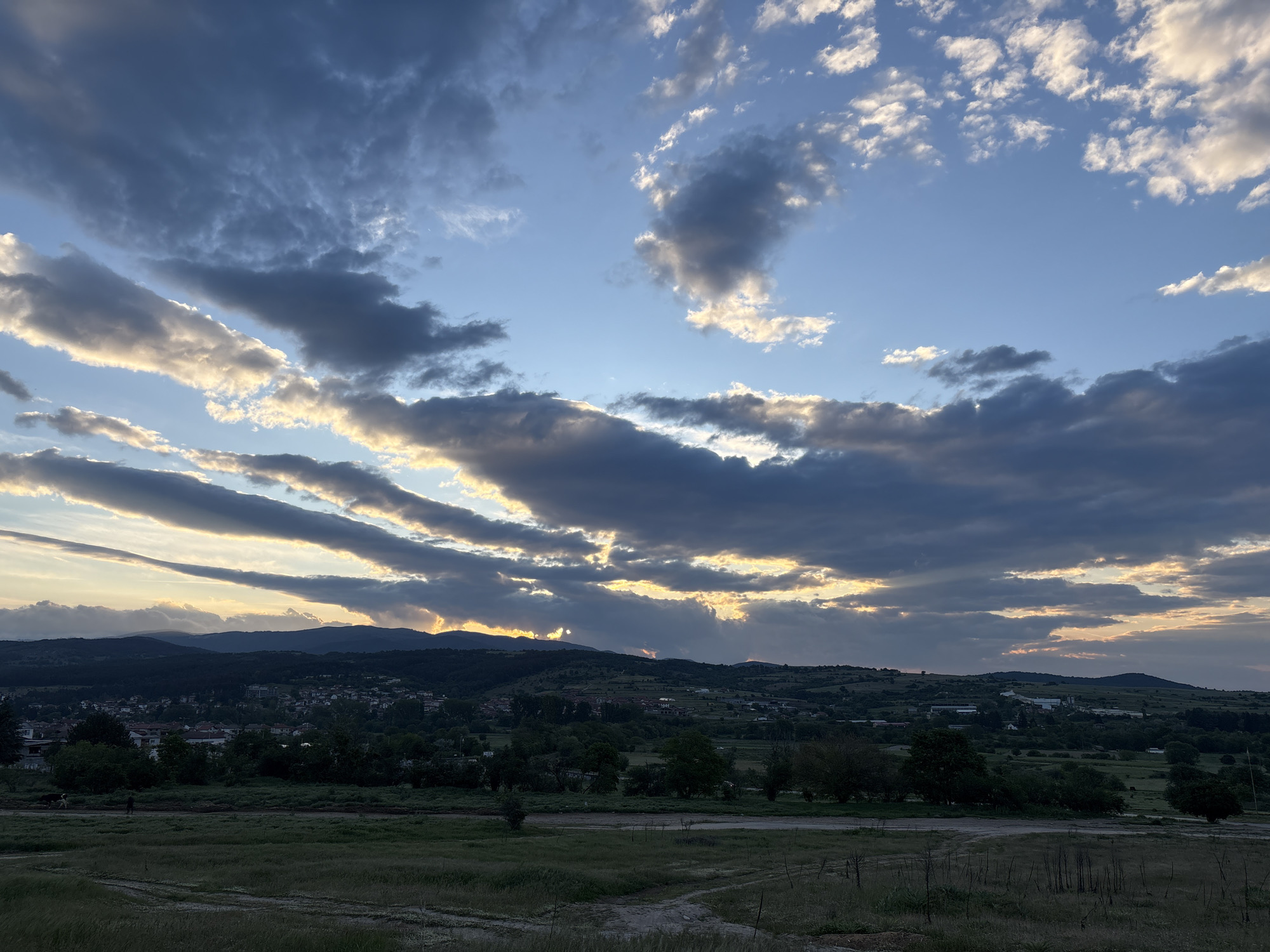 The image depicts a twilight or early morning scene in Strelcha, Bulgaria, characterized by a rural landscape under a dramatic sky. The foreground is dominated by a field of dry grasses, with visible dirt paths or tracks cutting through the vegetation. In the middle ground, a line of trees and shrubs separates the field from a distant vista of rolling hills and a portion of the town of Strelcha. Houses and buildings are discernible among the trees. The background features higher terrain, possibly mountains, silhouetted against the horizon. Above, the sky is filled with a mix of dark, heavy clouds and patches of blue sky. The clouds are illuminated from behind, suggesting the sun is either setting or rising, casting rays of light. The overall atmosphere is one of impending weather or the aftermath of a storm. No people are clearly visible in the image.