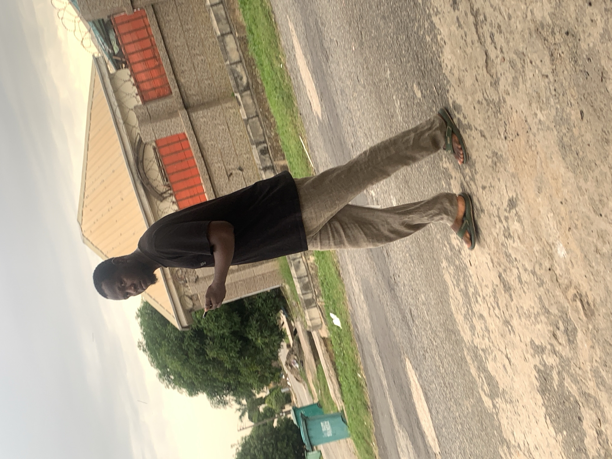 A Nigerian man wearing a black t-shirt and khaki pants walks on a paved road in Abuja, Nigeria. He is looking to his left and holding a mobile phone in his right hand. In the background, a beige building with a corrugated metal roof and red window grilles is visible, as well as lush green trees and two teal waste bins. The sky is overcast. The road is asphalt with some visible wear and tear, and patches of light-colored concrete.