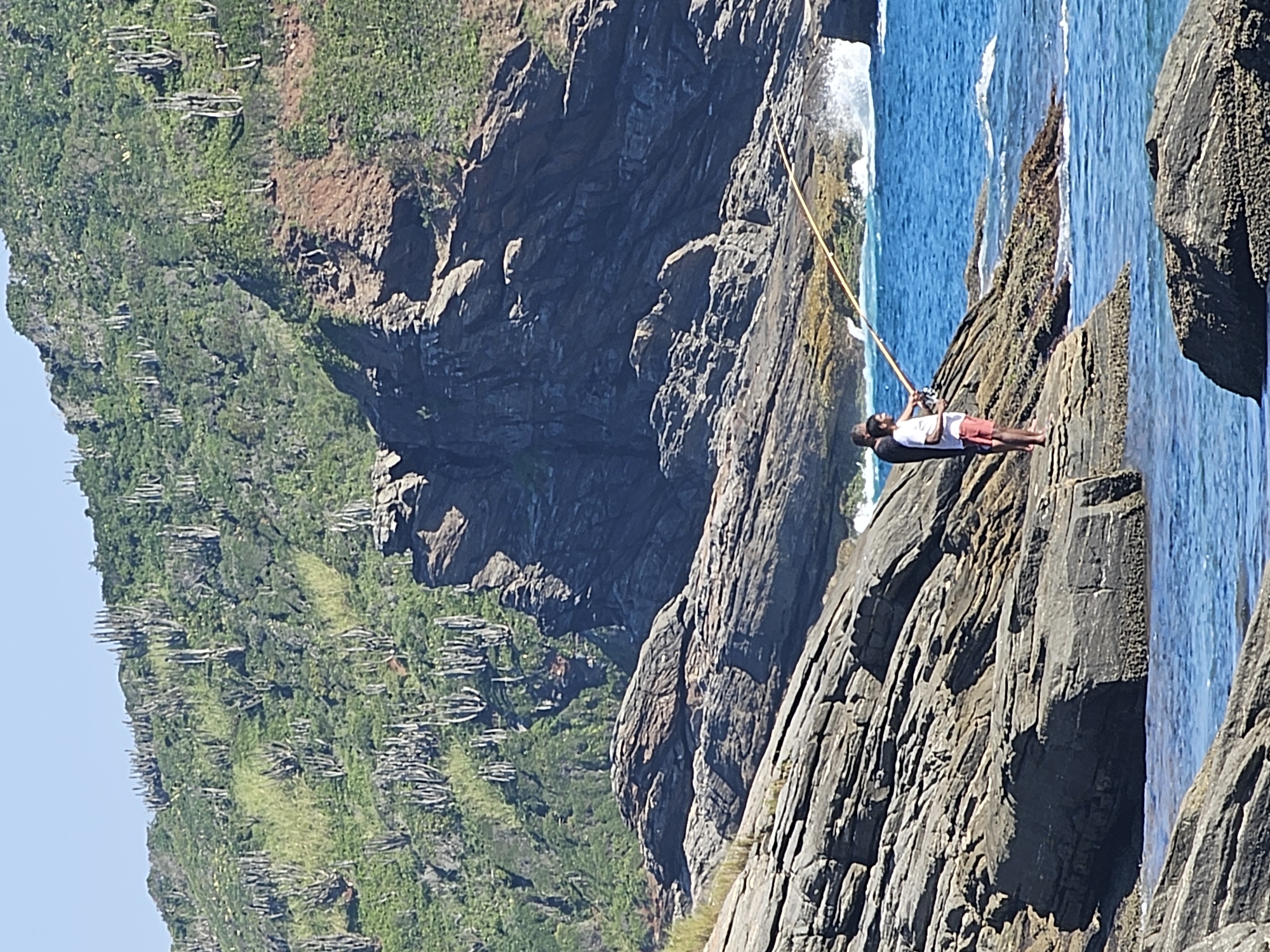 The image shows a man fishing off a rocky cliff in Armação dos Búzios, Brazil. The man, wearing a white shirt and red shorts, stands on the edge of the cliff holding a long, bamboo fishing rod. The cliff is composed of dark, greyish-brown rock formations. The ocean is a vibrant blue, with waves crashing against the rocks below. Lush green vegetation covers a hillside in the background, dotted with cacti or similar spiky plants.
