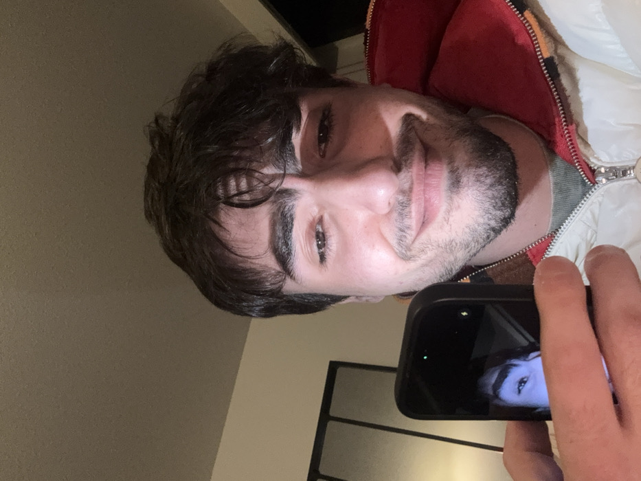 A fair-skinned man with dark hair and a trimmed beard holds a smartphone, taking a selfie. The phone screen reflects his eye. He wears a white and red jacket, partially zipped. The background is a neutral wall and a portion of a black metal frame. The image is taken at a slightly tilted angle.