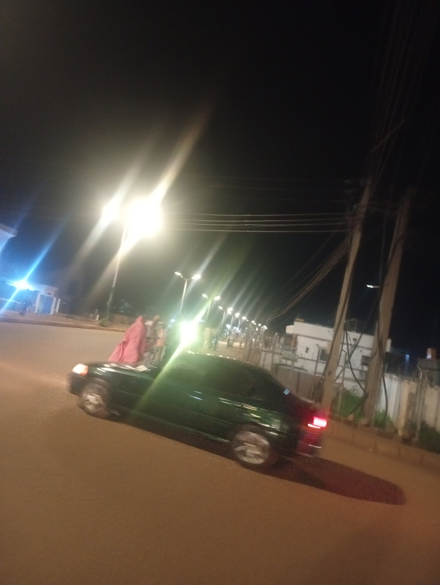 A dark green car is parked on a paved road at night in Bukuru, Nigeria. The car is facing right and its brake lights are illuminated. A person in a pink garment is standing next to the car. Streetlights are visible in the background, casting a bright glow. There are power lines and poles on the right side of the image. Buildings and a fence are also visible in the background.