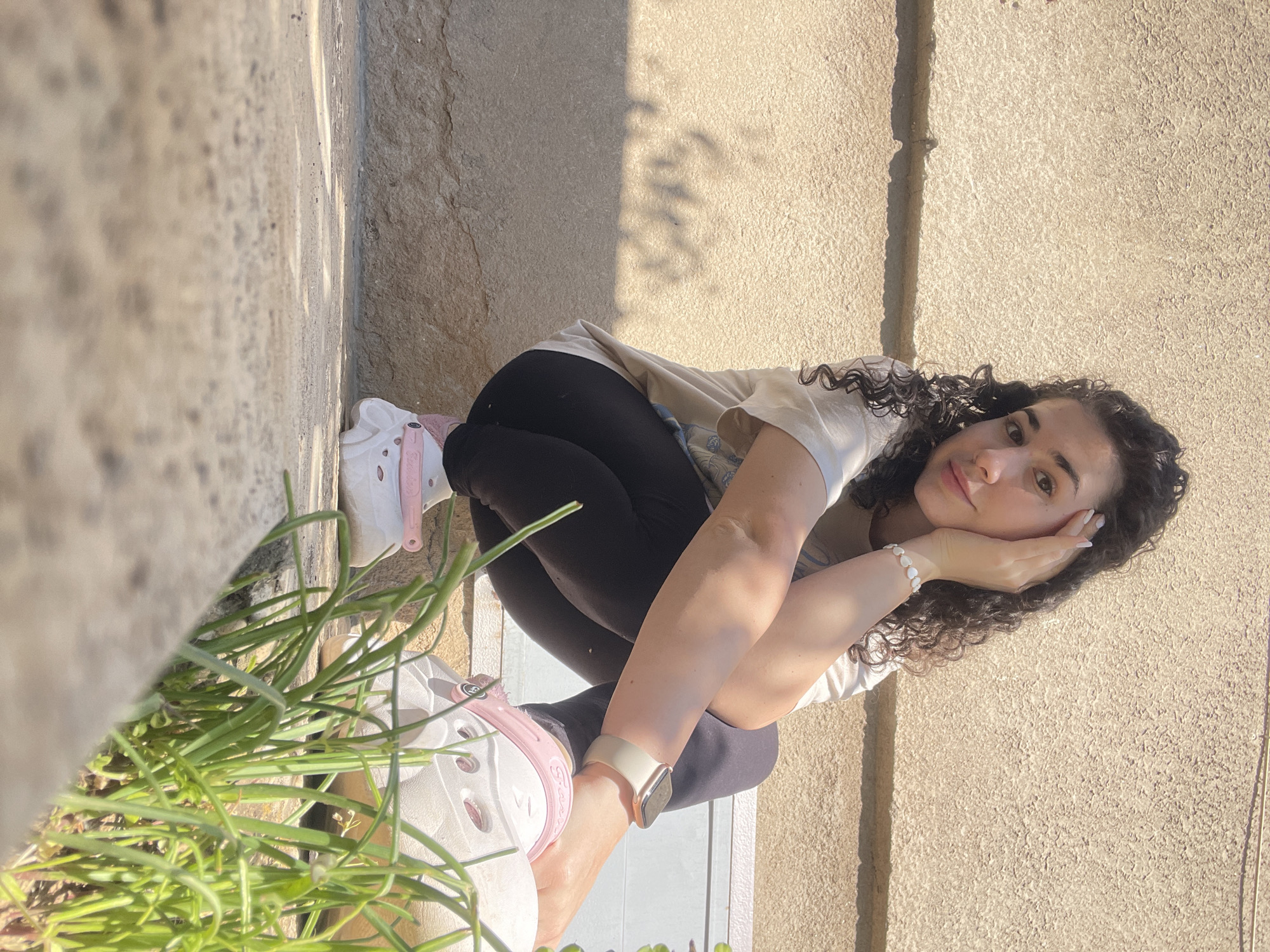 A young woman with curly dark hair is crouched on a concrete surface outdoors. She is wearing black leggings, a beige t-shirt, and white Crocs with pink details. She has a light-colored smartwatch on her left wrist and a bracelet on her right. She is resting her head on her hand and looking at the camera. In the lower left corner of the image, there are green plants. The background consists of a textured concrete wall and some lines in the concrete. The exact location in Sofia, Bulgaria, cannot be determined based on the visual information.