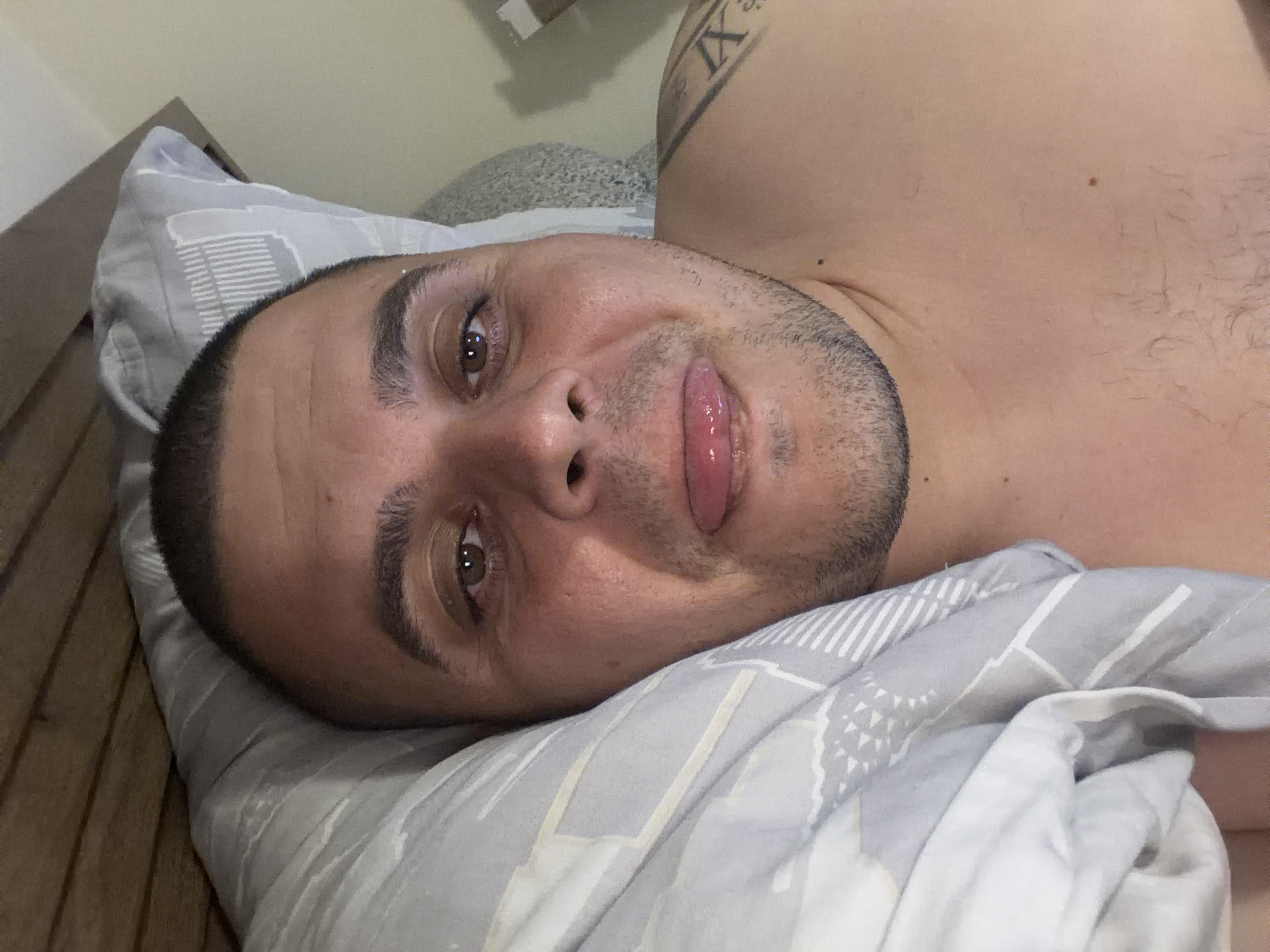 An adult man is depicted lying on his left side in what appears to be a bed, looking directly at the camera. His head rests on a light gray pillow with a white geometric pattern. He has short dark hair, a neatly trimmed stubble beard, and light brown eyes. His tongue is slightly extended, giving him a playful or cheeky expression. He is shirtless, and part of a tattoo featuring Roman numerals, specifically "IX", is visible on his right shoulder or upper arm.
He is covered partly by a light gray duvet or blanket, which also has a subtle pattern. To the left of his head, a wooden headboard or wall panel is visible, suggesting a bedroom setting. The overall lighting is soft and even, indicating an indoor environment, possibly during the daytime or with artificial light. The man's posture and facial expression convey a sense of relaxation and casualness. The image is taken from a slightly upward angle, with the man's head appearing on the left side of the frame. The location context provided is Shumen, Bulgaria.