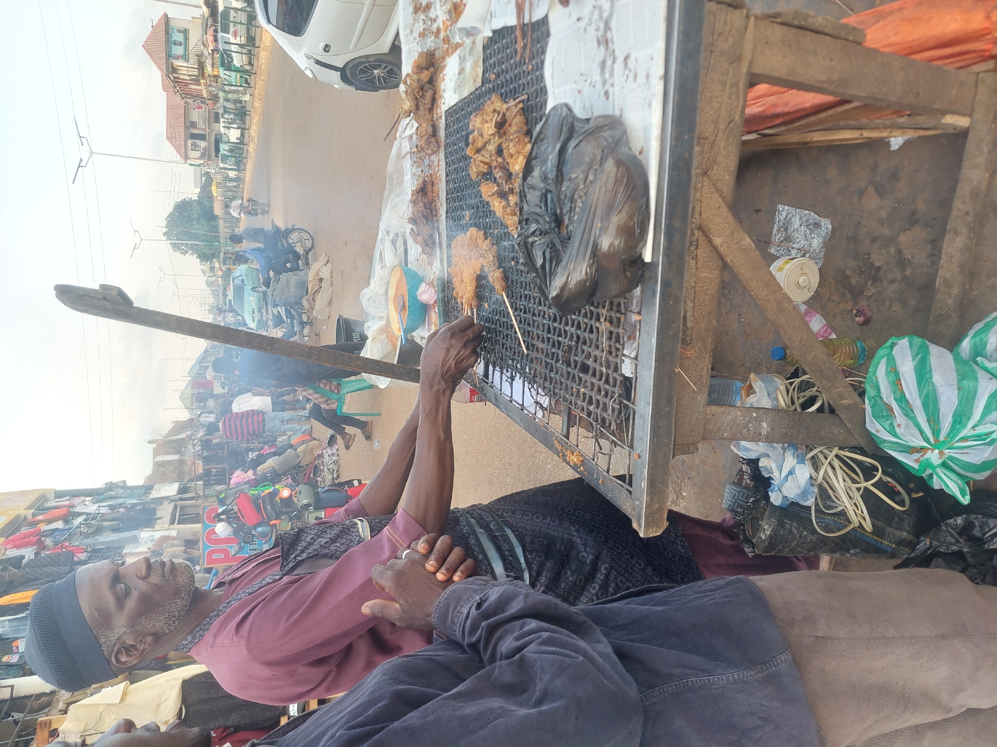 The image depicts an outdoor street scene in an unknown city or town, featuring a street food vendor grilling meat.
In the foreground, a dark-skinned man with a salt-and-pepper beard and a dark cap stands behind a metal grill table. He wears a maroon long-sleeved shirt and a dark patterned apron/wrap. His right hand holds a skewer with meat over the grill, while his left arm is extended towards the cooking area. A small stick or toothpick is in his mouth. To his immediate left, partially visible, another dark-skinned man in a dark blue long-sleeved shirt and light brown pants stands with his hands clasped, observing.
The grill table features a wire mesh top, supported by wooden legs. On the grill, multiple skewers of meat and loose pieces of meat are cooking, some coated with a reddish spice mixture. A black plastic bag rests directly on the grill surface. To the left of the cooking area, a light blue bowl contains a reddish sauce and a sliced red onion. Below the table, the ground is unpaved, showing dirt and scattered debris including plastic bags (one green and white striped, others black), coiled white electrical wires, and discarded plastic bottles. A tall, rough-hewn wooden pole stands vertically on the far left.
The background reveals a bustling street scene. A paved road stretches into the distance, lined with streetlights that appear to have solar panels. Vehicles, including motorcycles and a white car (possibly a small hatchback), are present on the road. Buildings line both sides of the street; the structure on the left displays clothing for sale, suggesting market stalls, while a building on the right has a reddish-brown roof and posters on its exterior. Numerous people are visible in the midground and background, walking, sitting, or riding vehicles, indicating a lively commercial area. The sky is a bright, hazy white, and subtle smoke rises from the grill.