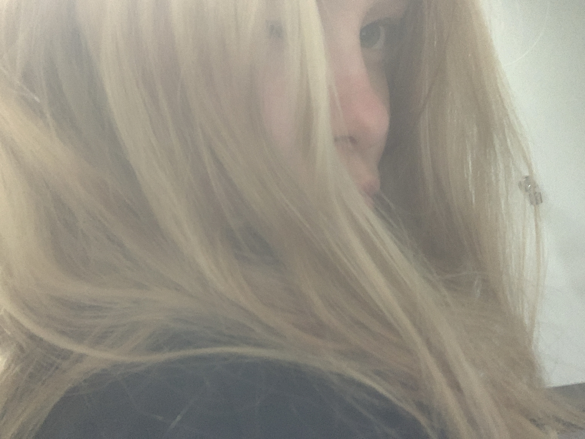 The image is a close-up, slightly out-of-focus, medium shot of a person's face partially obscured by their blonde hair. The person is looking to their right. The visible features include an eye, part of a nose, and lips. The lighting is soft and diffused, creating a hazy effect. The background is a plain, light color, likely a wall. There is no indication of the city of Sinyavino, Russia, or any specific environmental details that would place the image in that location. The focus is entirely on the facial features and hair.