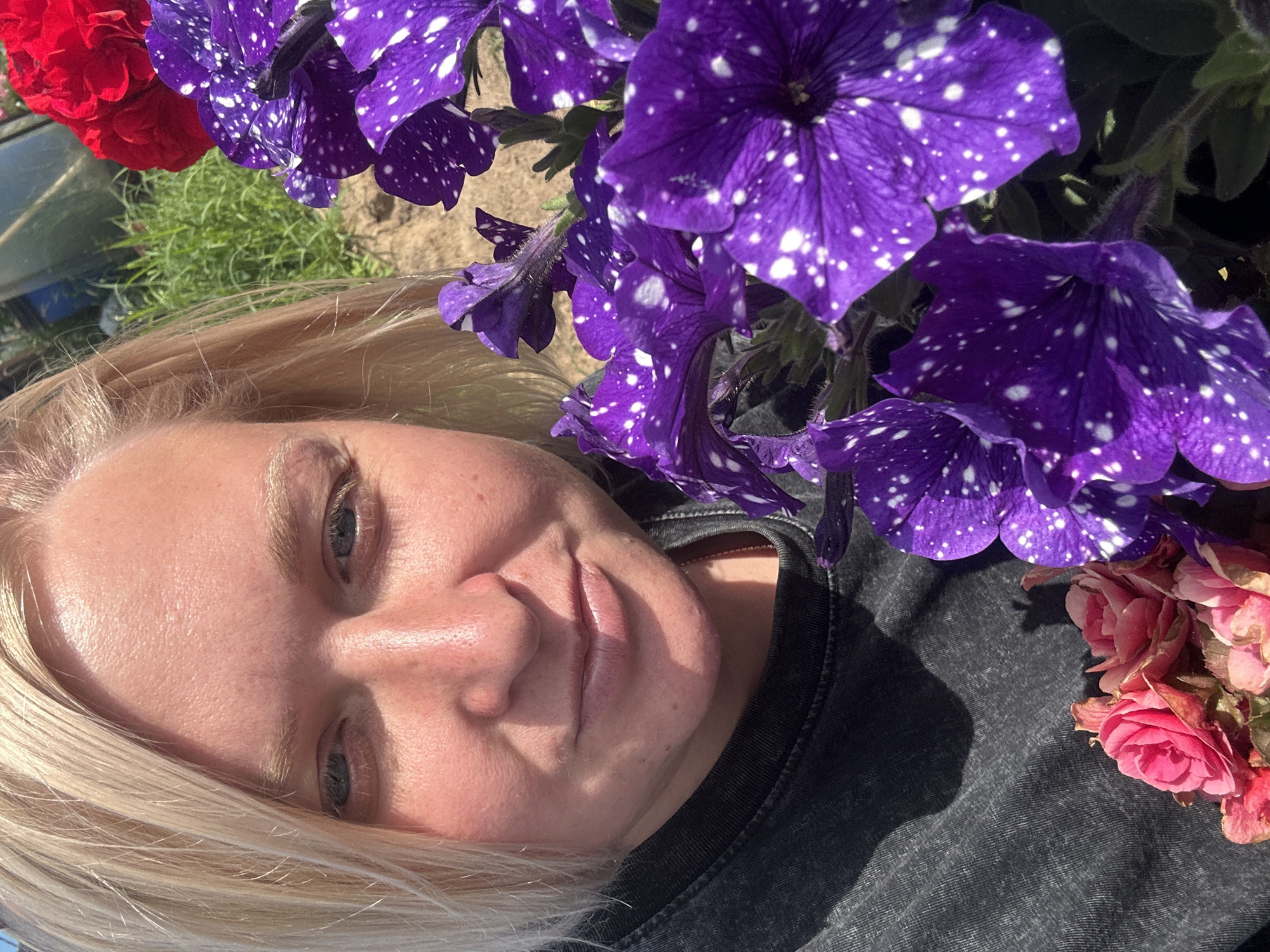 In a close-up, outdoor selfie, a blonde woman with blue eyes and fair skin is partially visible. She is wearing a dark grey t-shirt. To her right are several vibrant purple petunias with white speckles, and to her left, a cluster of small pink flowers. In the background, out of focus, is green foliage and hints of structures, possibly a greenhouse, under a bright sky. The image appears to be taken in Chaikovsky, Russia.