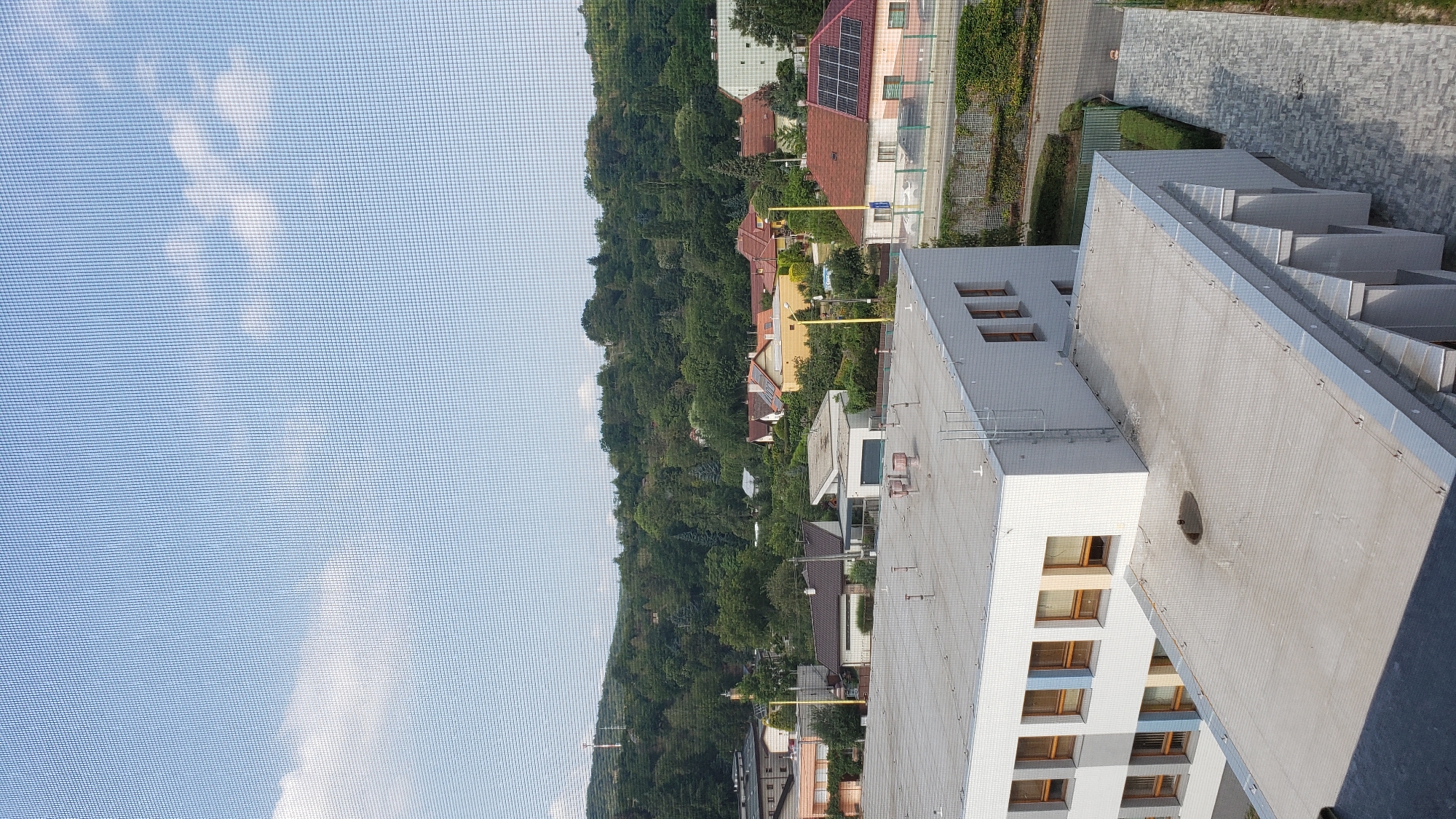 The image presents an aerial view of a suburban or semi-urban area, likely Prešov, Slovakia, looking towards a densely wooded hillside. In the foreground, the flat roofs of modern buildings are visible. The building closest to the camera is white with multiple floors, featuring rows of windows with wooden frames. Adjacent to it, another building with a flat, grey roof has several ventilation pipes and a ladder attached to its side. A second, lower flat roof is in the center, also grey, with a dark circular vent.
Beyond these buildings, a scattering of houses with pitched roofs, predominantly in shades of orange, yellow, and white, are nestled amongst lush green trees. Some houses have solar panels on their roofs, suggesting a contemporary residential area. A prominent feature is a tall, yellow pole visible amongst the houses. The background is dominated by a verdant, tree-covered hill, which rises towards a pale blue sky with scattered white clouds. A slender, red-and-white telecommunications tower is visible on the crest of the hill in the distance. The overall impression is a scene of developed residential areas integrated with natural, forested landscapes.