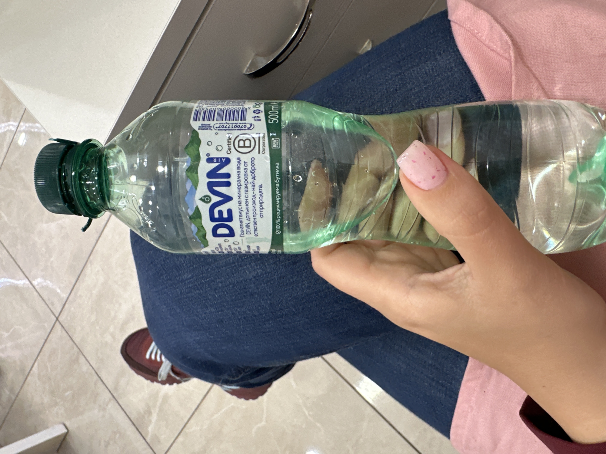 A person wearing blue jeans, a pink shirt, and red and white sneakers is holding a 500ml bottle of Devin mineral water. The bottle has a green cap and a label with the Devin logo and Bulgarian text. The person's hand is visible, with a light pink fingernail. The background shows a tile floor and a cabinet with a silver handle. The scene appears to be indoors, possibly in a commercial setting like a salon or waiting area. There is no clear evidence indicating the location is in Sofia, Bulgaria, beyond the Bulgarian text on the water bottle label.
